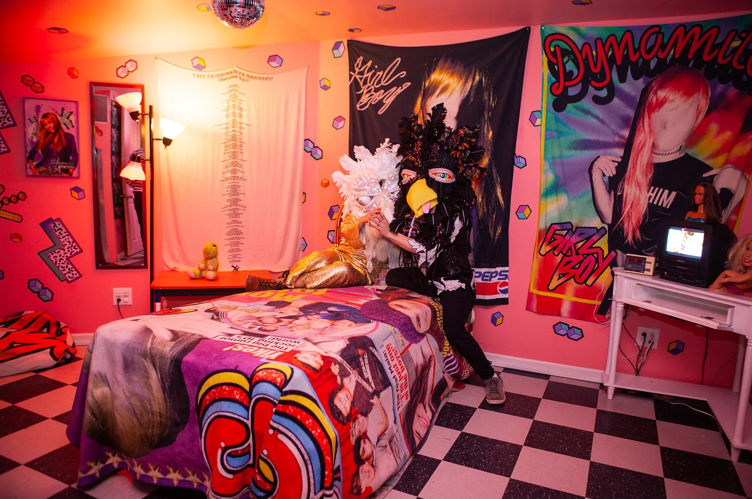 A pink themed room with posters and artwork, featuring two people in elaborate costume masks dancing by a bed with a wrap featuring colorful pop art. The room has checkered black and white flooring, a small TV, a mirror, and wall decorations.