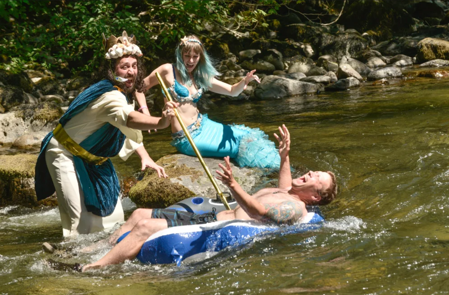 Three people dressed as characters, two women in mermaid costumes and one man in a pirate costume, helping a man on a raft in a river with rocks and trees in the background.