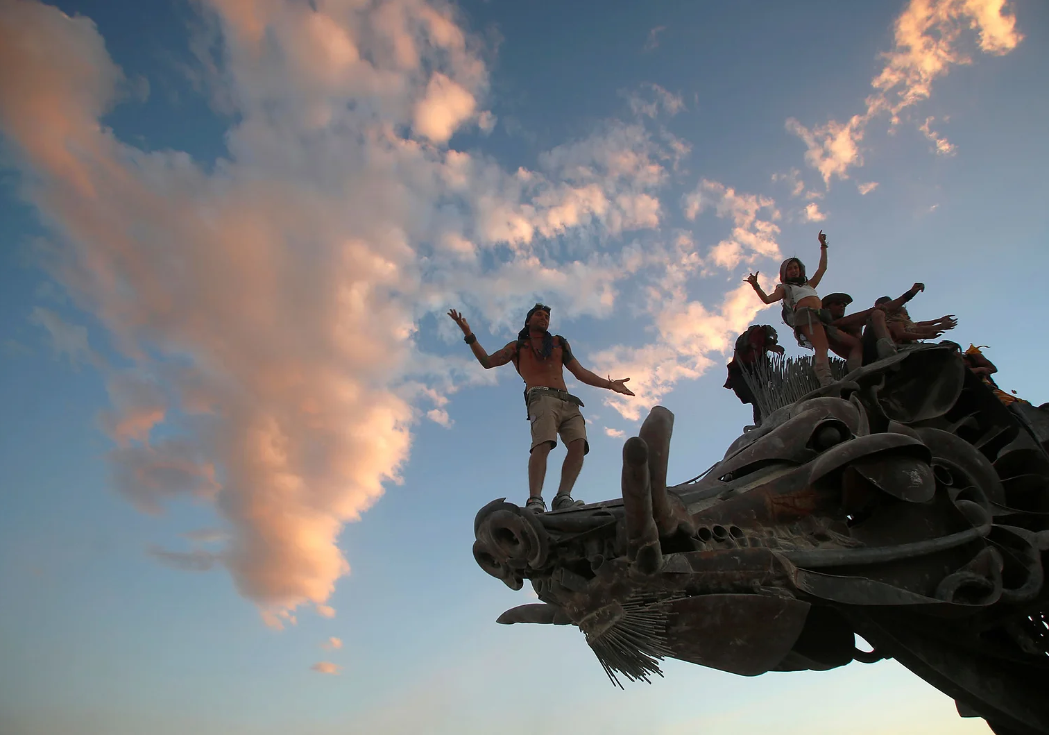 People standing on a creative metallic sculpture resembling a vehicle or animal, with a partly cloudy sky at sunset in the background.