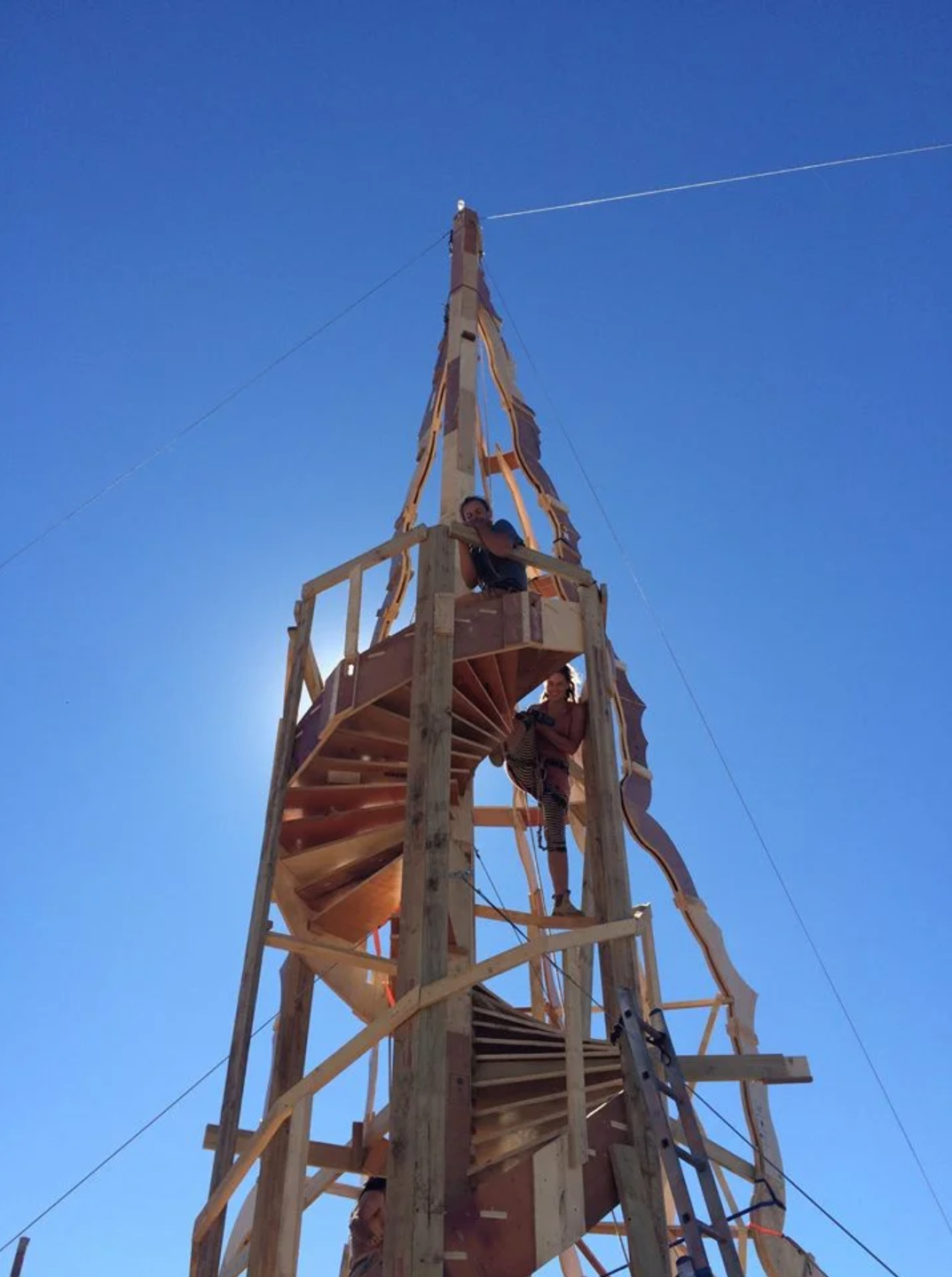 People building a tall wooden spiral staircase structure outdoors under a clear blue sky.