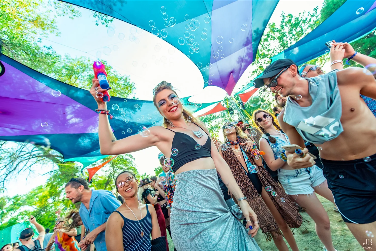 People enjoying a music festival outdoors, dancing and smiling under colorful canopy drapes, with some wearing sunglasses and casual summer clothing.