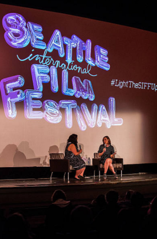Two women sitting on stage in conversation at the Seattle International Film Festival, with a large screen behind them displaying its logo and the hashtag #LightTheSIFFUpdate.