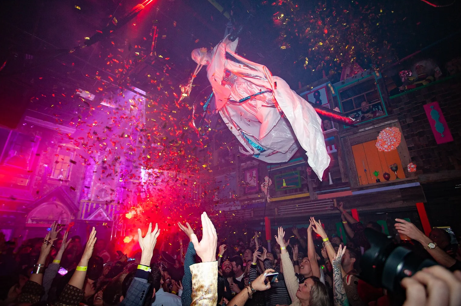 People reaching towards a flying unicorn piñata as pink and gold confetti falls down inside a lively bar or club with colorful decorations.