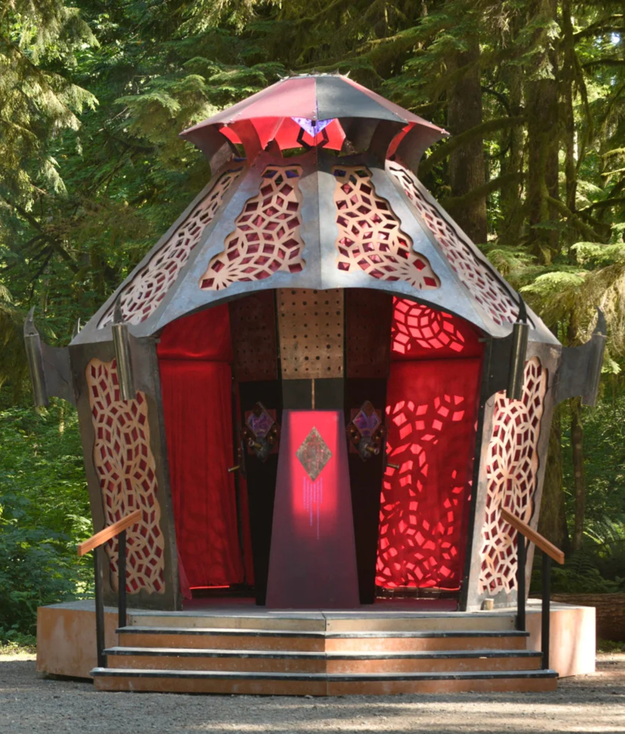 An ornate structure resembling a lantern with intricate cut-out patterns, surrounded by a forest of tall green trees.