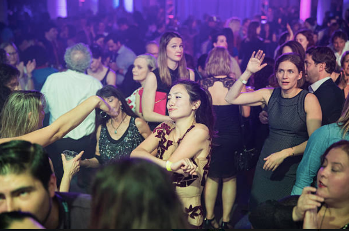 Crowded dance floor at a nightclub with people dancing and socializing under colorful, purple and blue lighting.