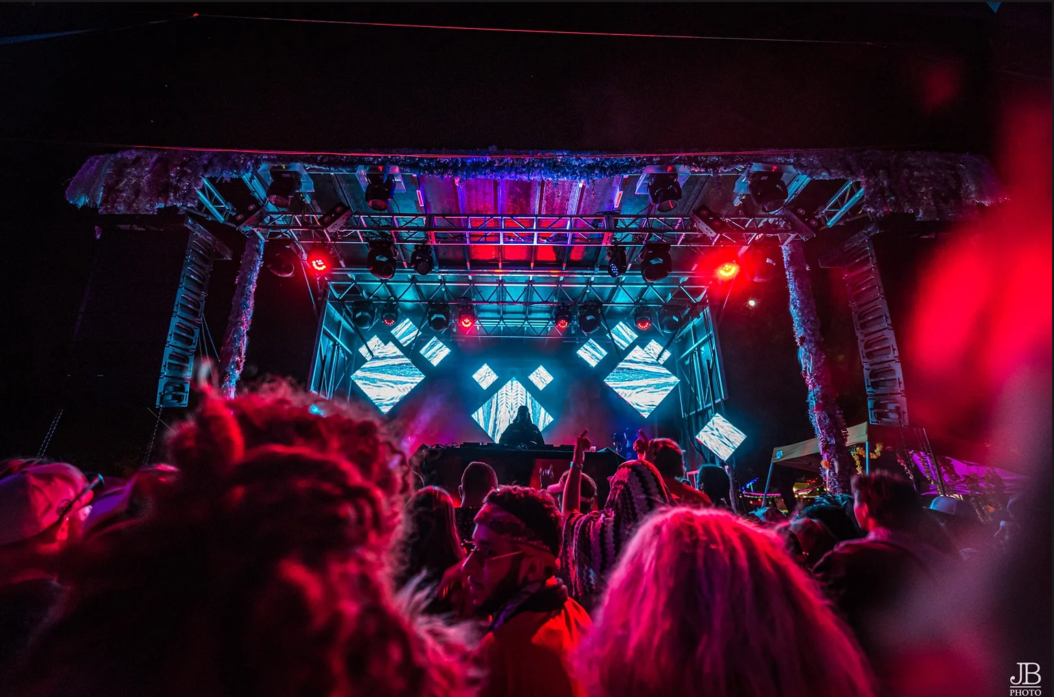 Nighttime concert scene with a large crowd facing a brightly lit stage featuring LED screens, stage lights, and a DJ or performer silhouette in front of the screens.