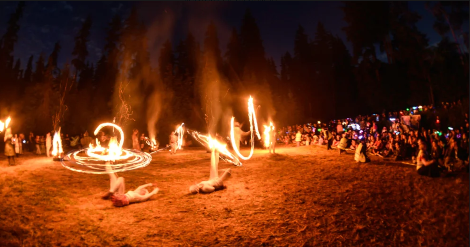 People performing fire poi spinning at night in an outdoor forest setting with many spectators watching.