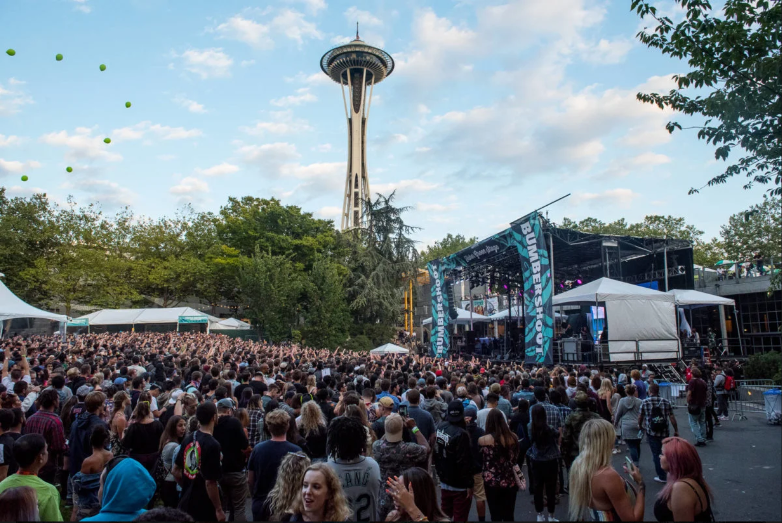 Crowd attending an outdoor concert near the Space Needle in Seattle, Washington, with a stage, tents, and balloons in the sky.