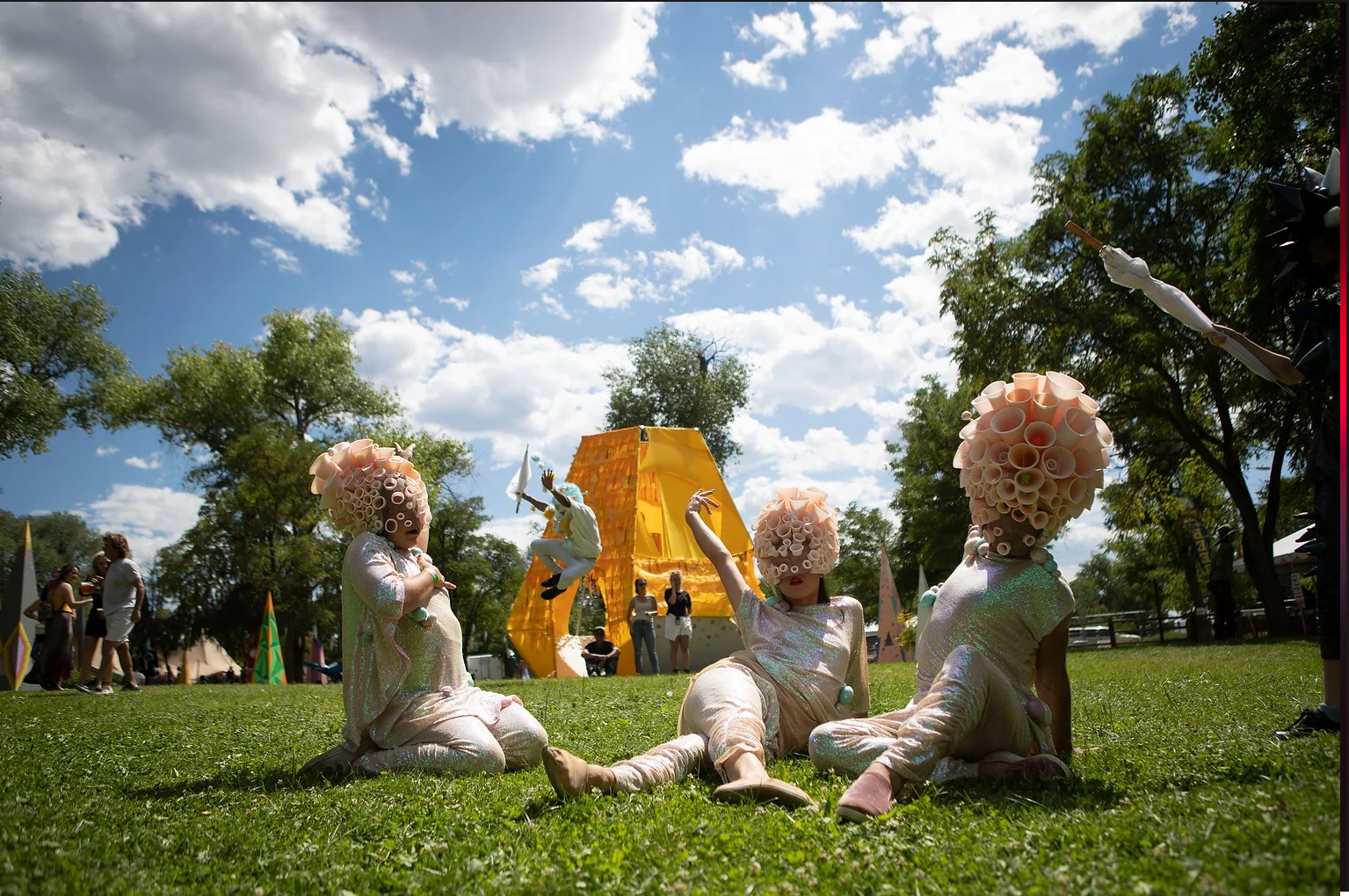 Three performers dressed in elaborate costumes with large pink bubble headpieces sitting on grass at an outdoor event, with a yellow tent structure and trees in the background under a partly cloudy sky.