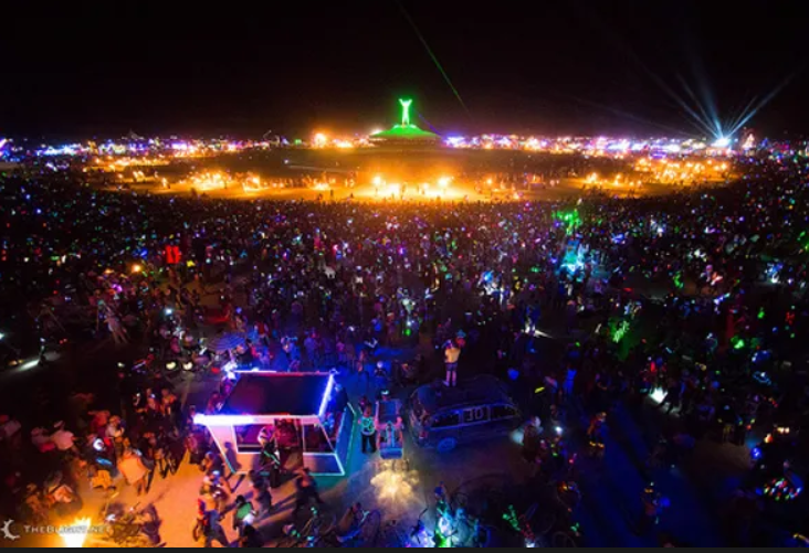 Nighttime scene of a large outdoor festival with many people illuminated by colorful lights, and a prominent green glowing sculpture or monument in the distance.