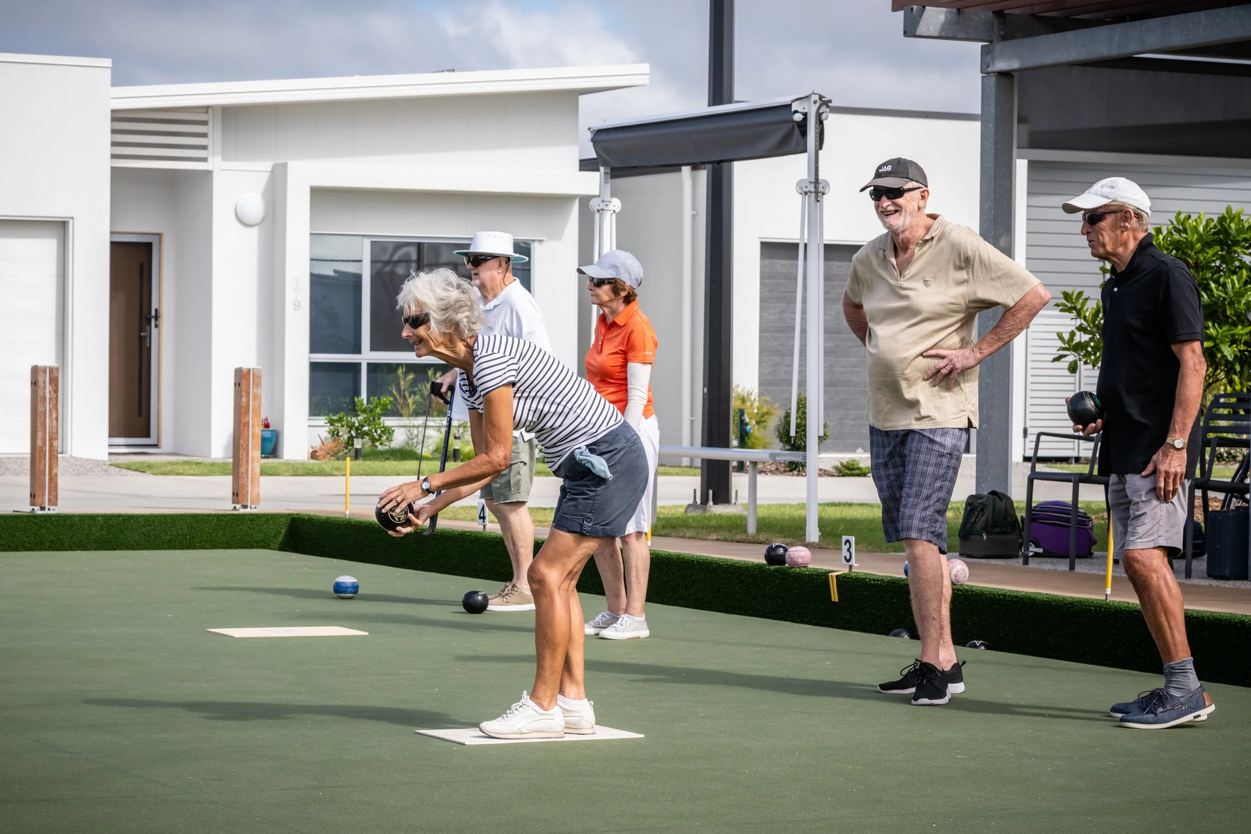 Group of senior people playing bocce ball on a green court outdoors, with modern houses in the background.