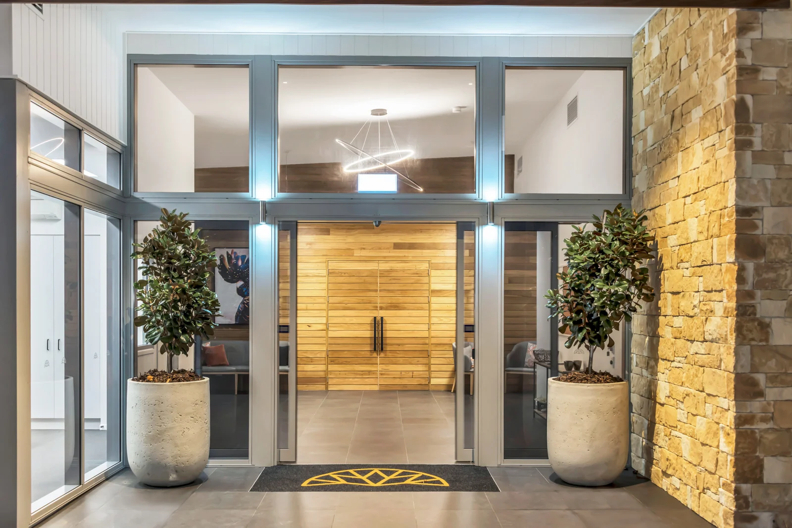 Modern entrance with large glass doors, potted plants, stone wall, and wooden accents inside.