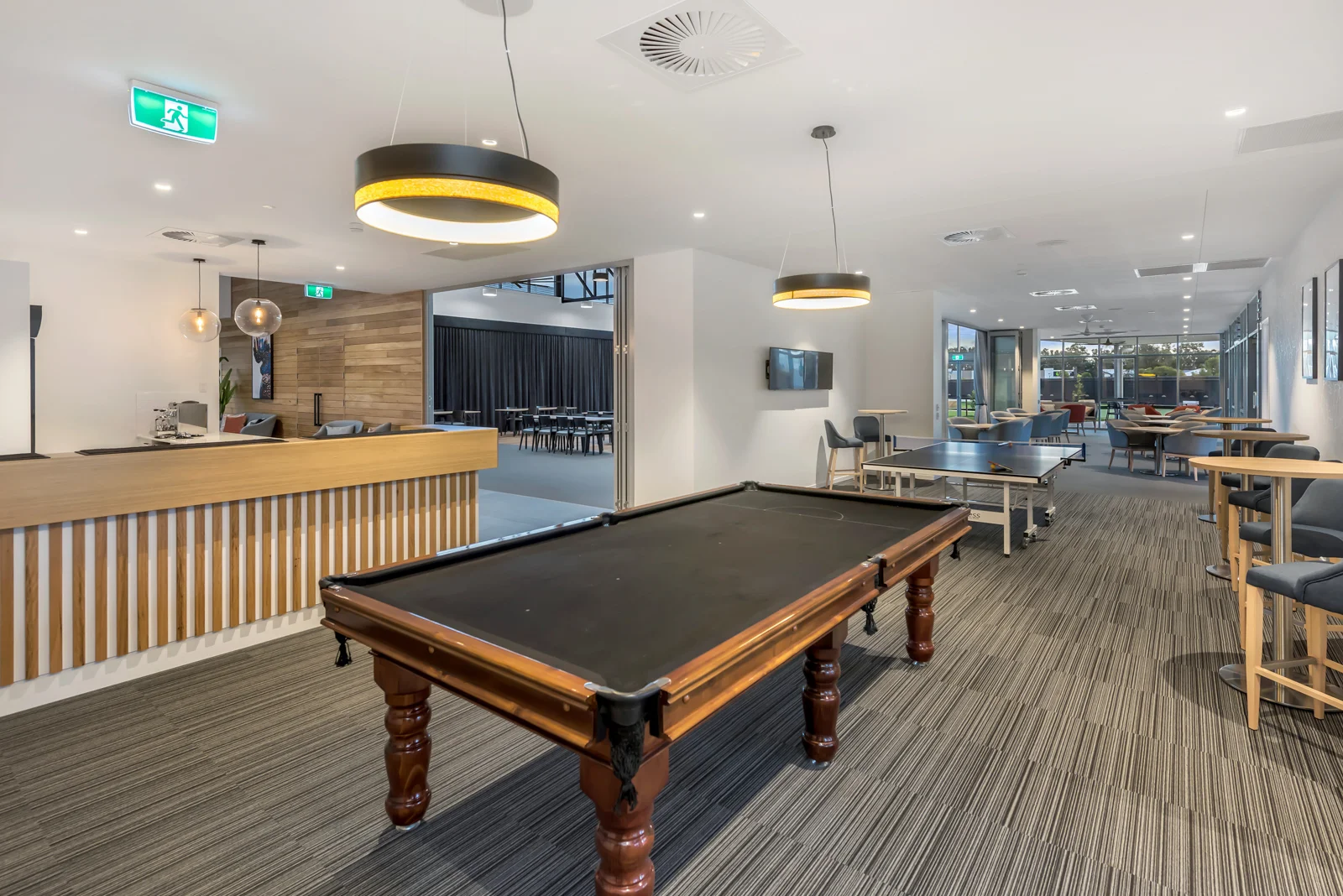 Indoor recreational area with a pool table, high tables with chairs, and a dining or lounge space with multiple tables and chairs. Large windows let in natural light.