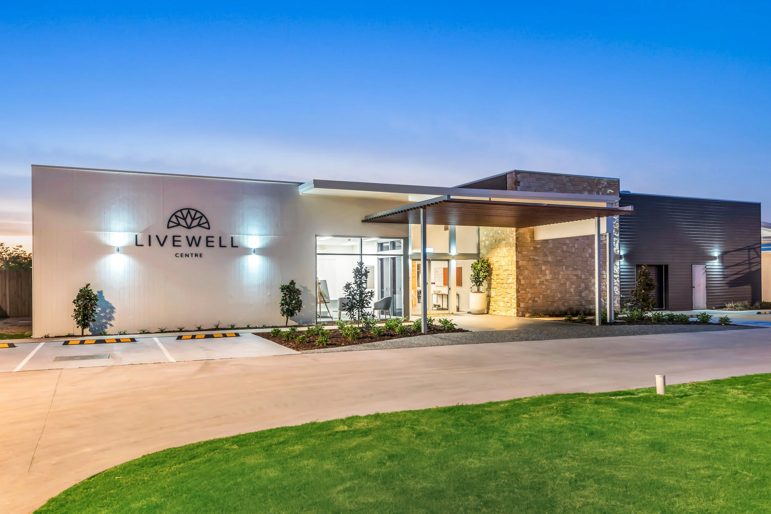 Exterior view of the Livwell Centre building at dusk, featuring a modern design with a white wall, glass entrance, and landscaping with small trees and plants.