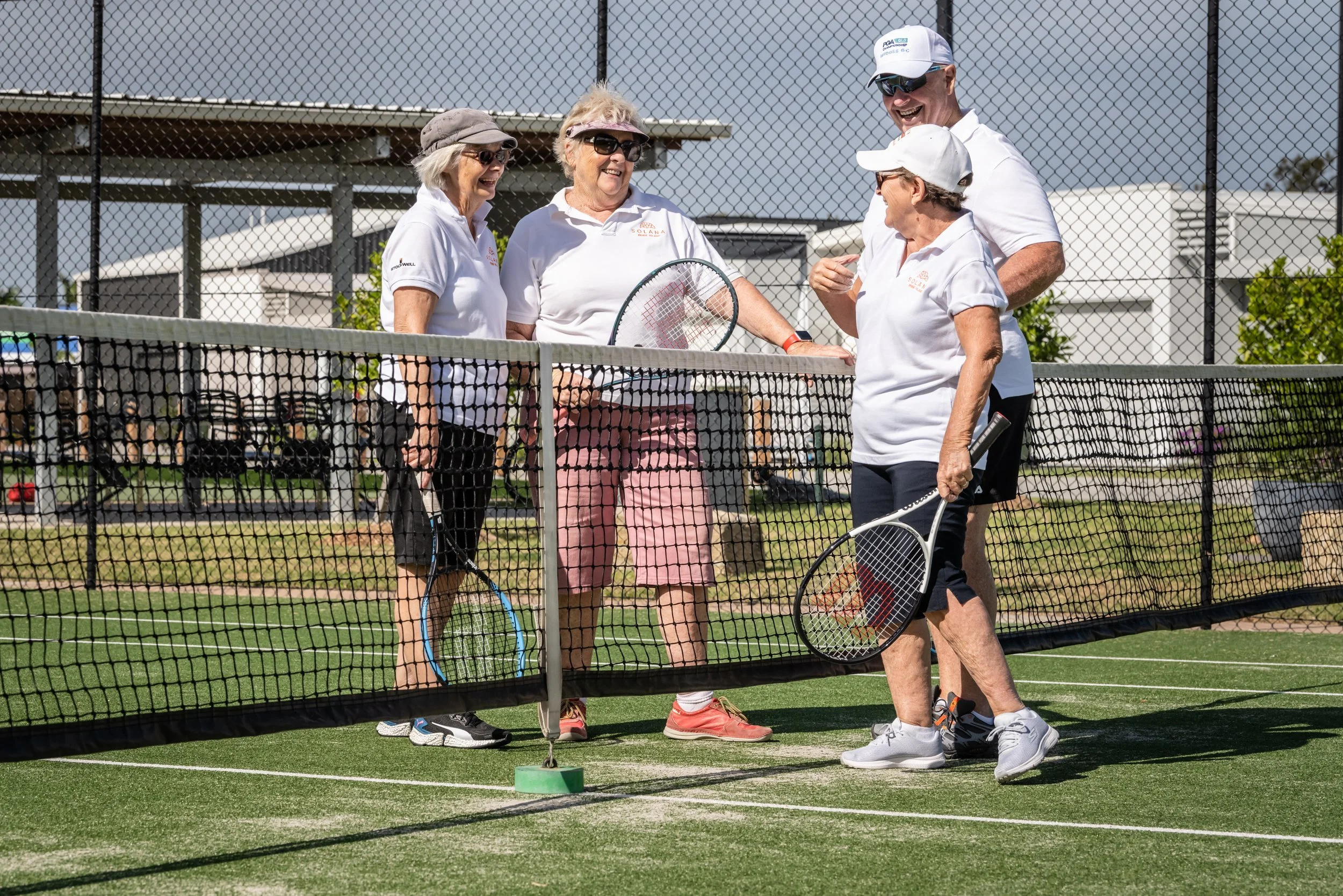 Group of five seniors playing pickleball on an outdoor court, smiling and talking, holding paddles, with a chain-link fence and buildings in the background.