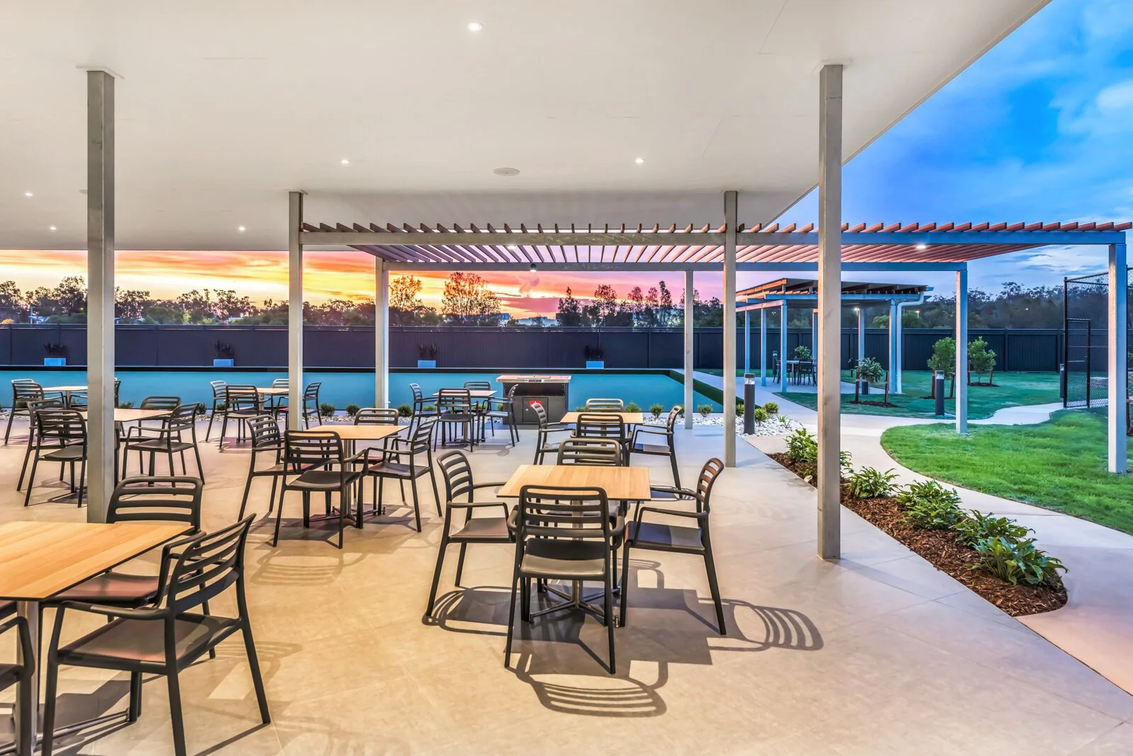 An outdoor patio area with wooden tables and black chairs, green lawn, and a swimming pool, with the sunset sky visible in the background.