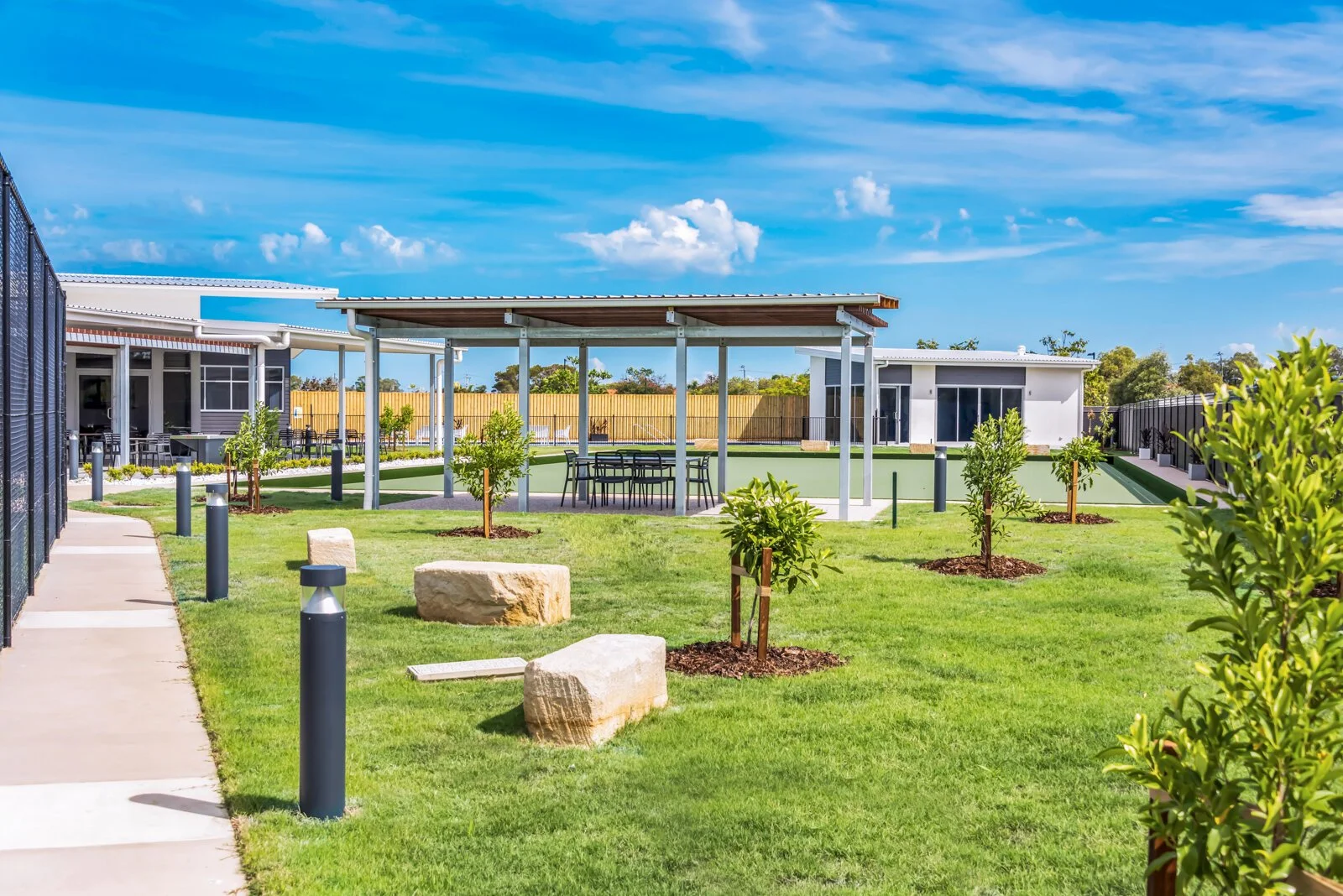 Outdoor area with green grass, young trees, stone benches, and a shaded seating area, surrounded by modern white buildings on a sunny day with a blue sky and some clouds.