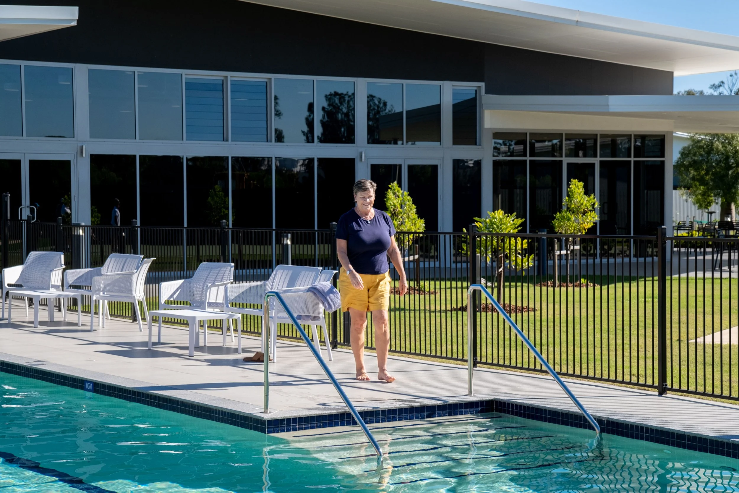 A woman in a navy T-shirt and yellow shorts walking barefoot by a swimming pool. The pool area has white chairs and a black fence, with a modern building and green trees in the background.
