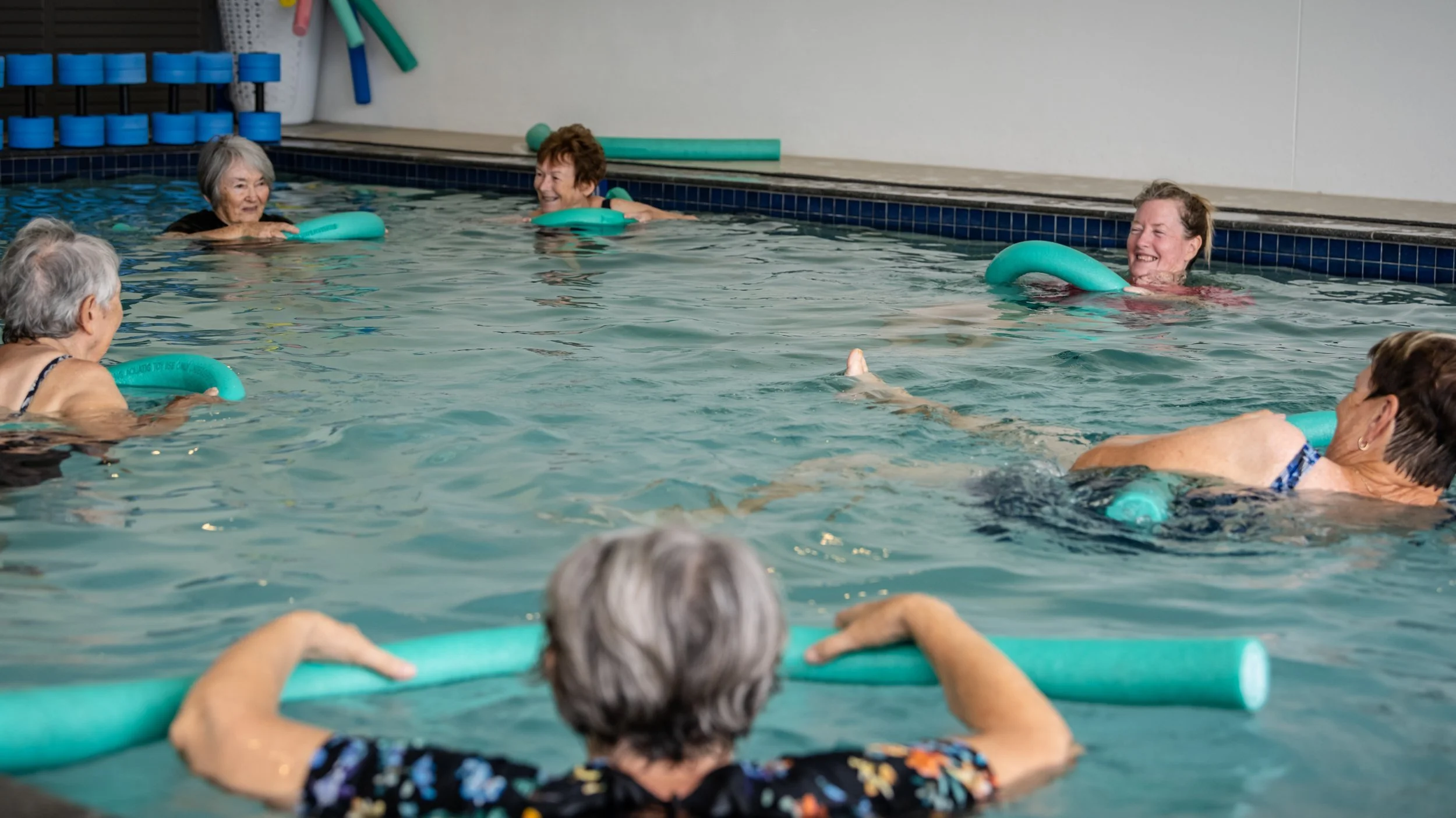 Group of elderly women participating in a water exercise class in a swimming pool, holding pool noodles and smiling.