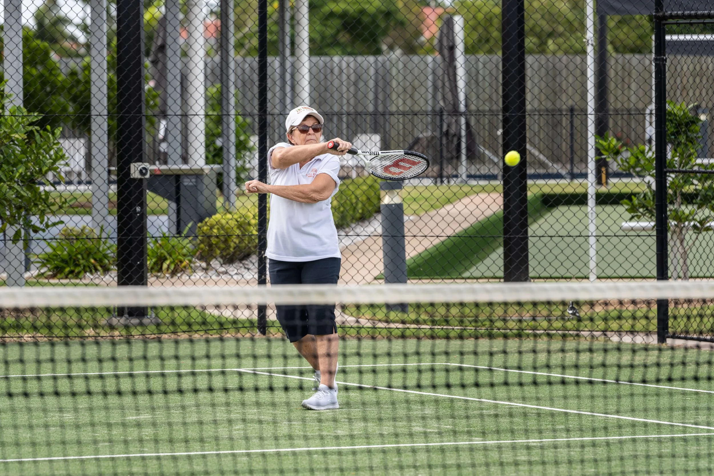 An older woman playing pickleball on a court, hitting the ball with a paddle, wearing sunglasses, a white shirt, and dark shorts, behind a chain-link fence.