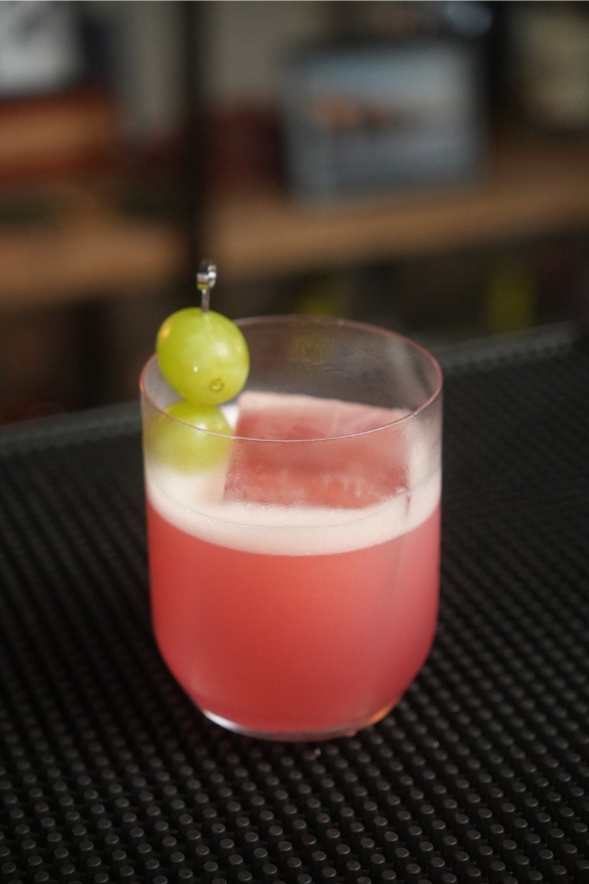 A savory enzoni pink cocktail in a glass garnished with two green grapes on a skewer.