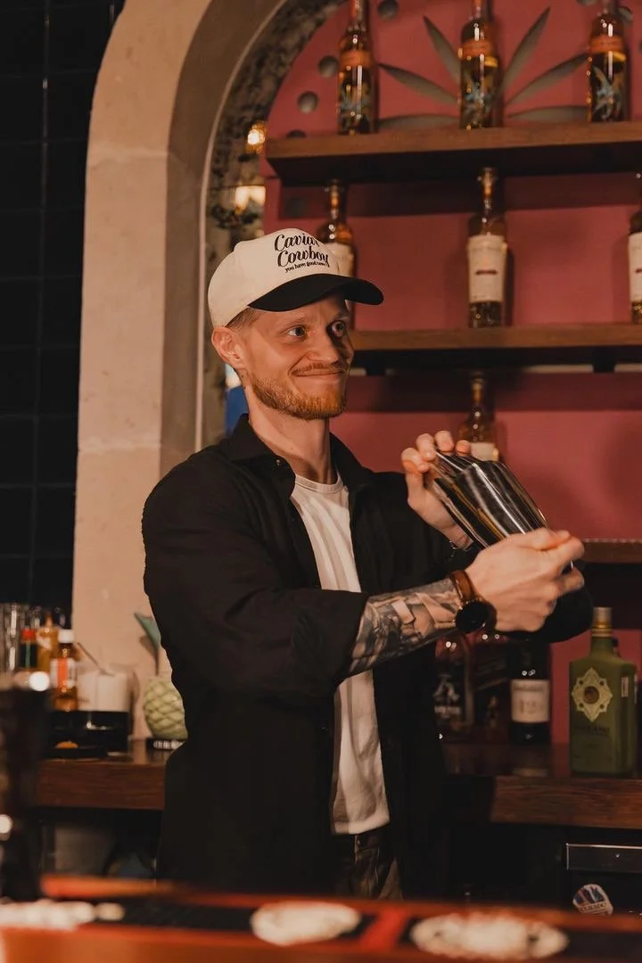A man with a beard and tattooed arm mixing a cocktail in a bar with pink walls and bottles on shelves behind him.