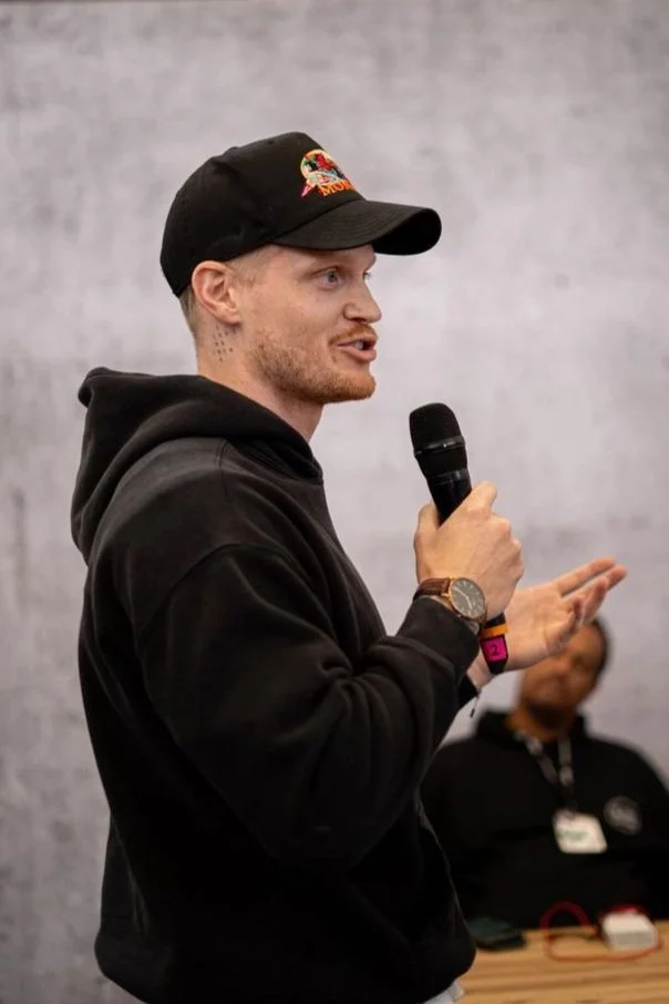 Chris, Not Just a Bartender, with a black cap, black hoodie, and a watch is speaking into a microphone at a bartending event, with a gray wall in the background.