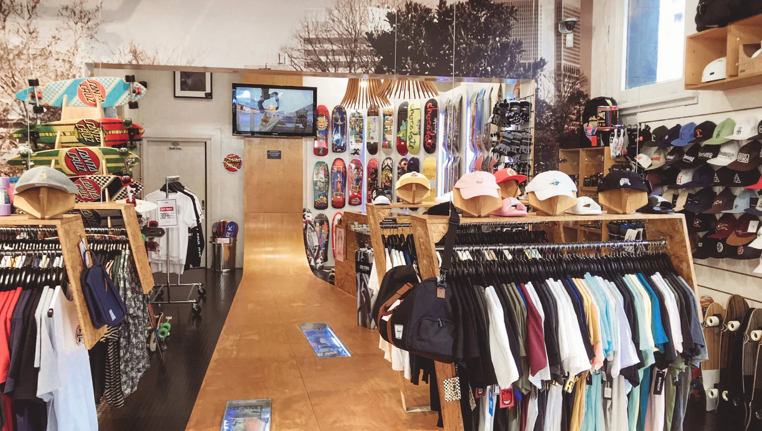 Inside a retail store displaying skateboards, caps, t-shirts, and accessories with a mountain mural wall and a TV screen showing a skateboarding video.