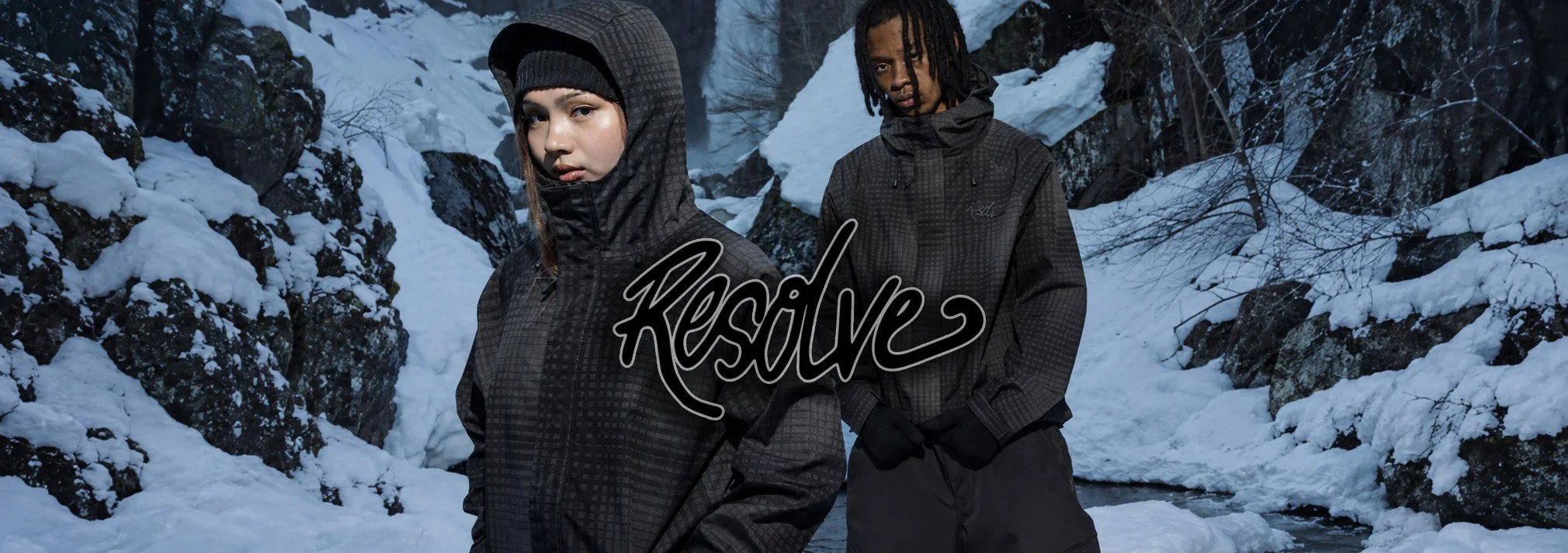 Two young people in black patterned winter jackets and gloves standing in a snowy, rocky outdoor environment with snow-covered trees and a partially frozen stream, with the word 'Resilence' written across the image.