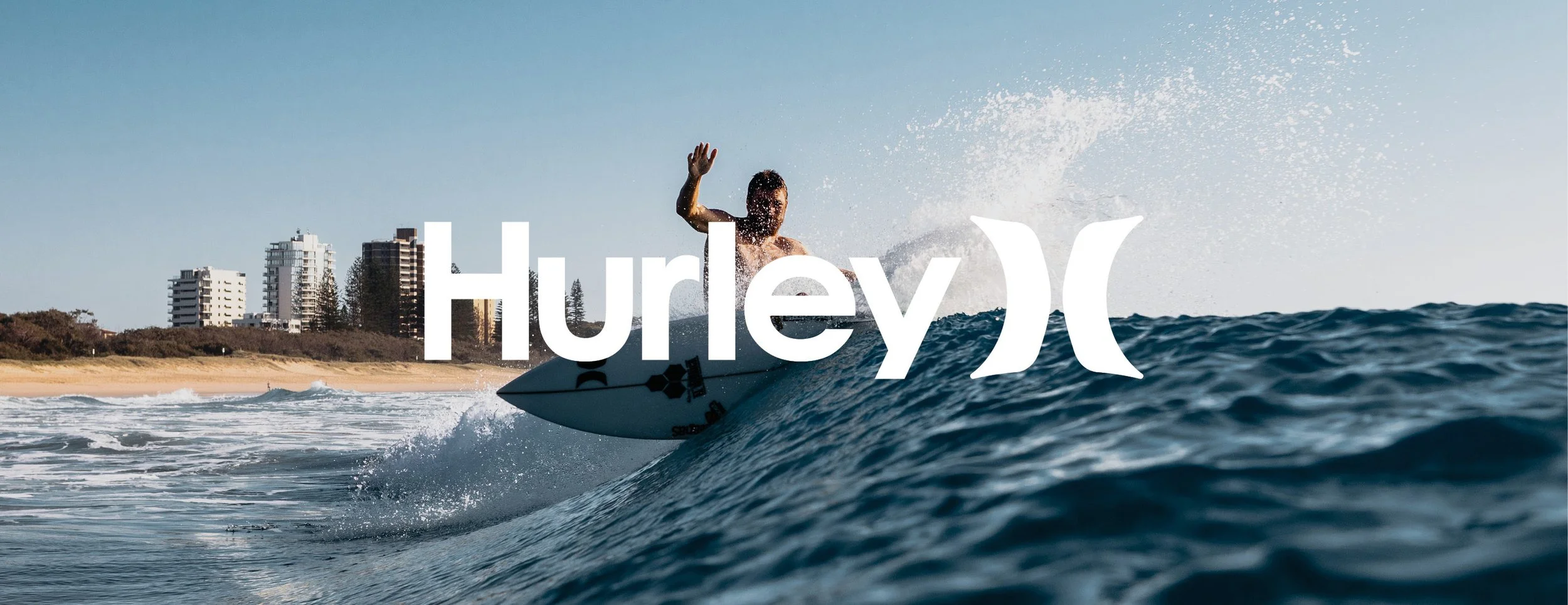 A surfer riding a wave near the beach with buildings in the background, with the Hurley logo overlaid.