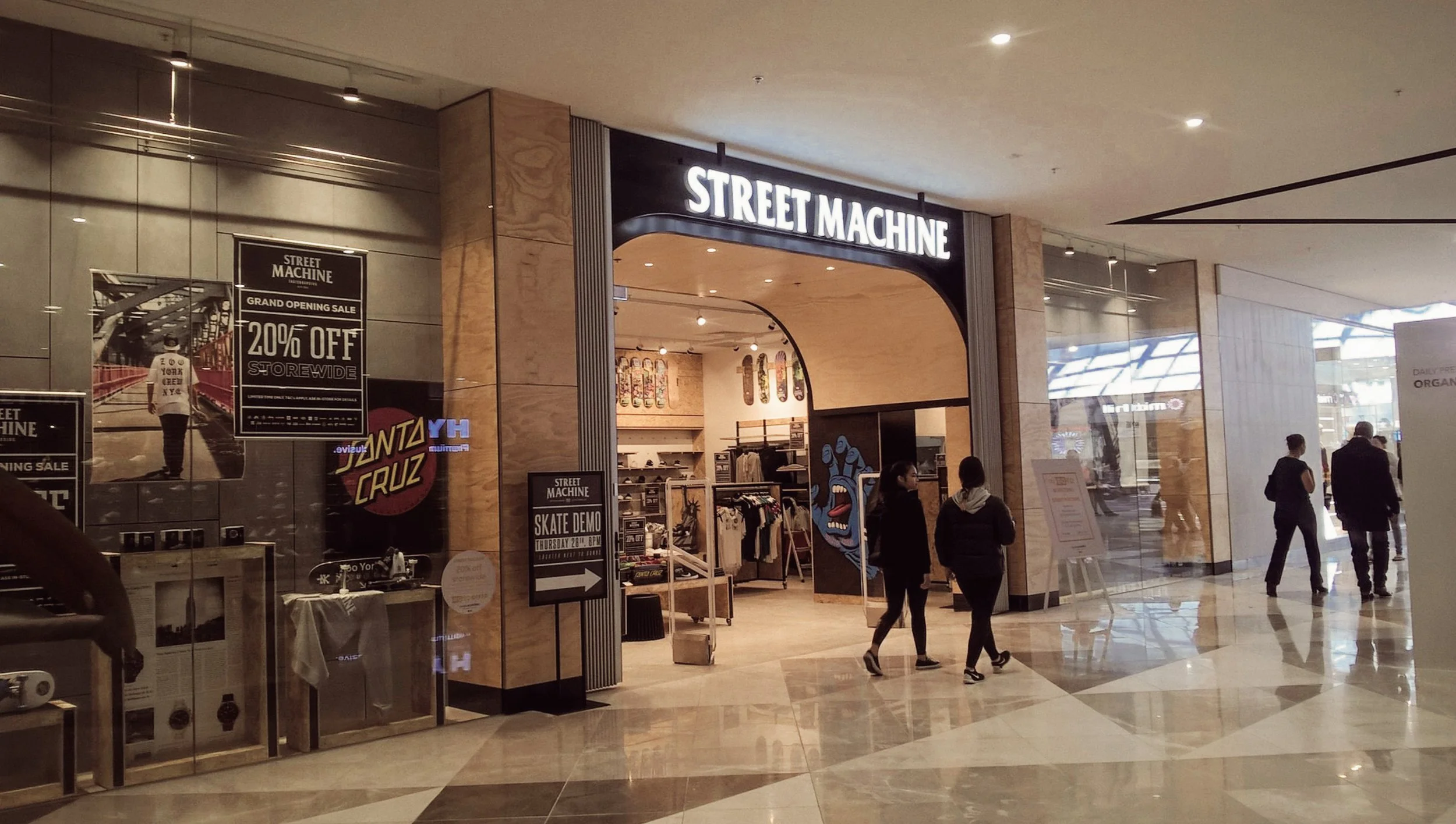 Entrance to STREET MACHINE retail store in a shopping mall, with shoppers walking by and promotional posters on display.