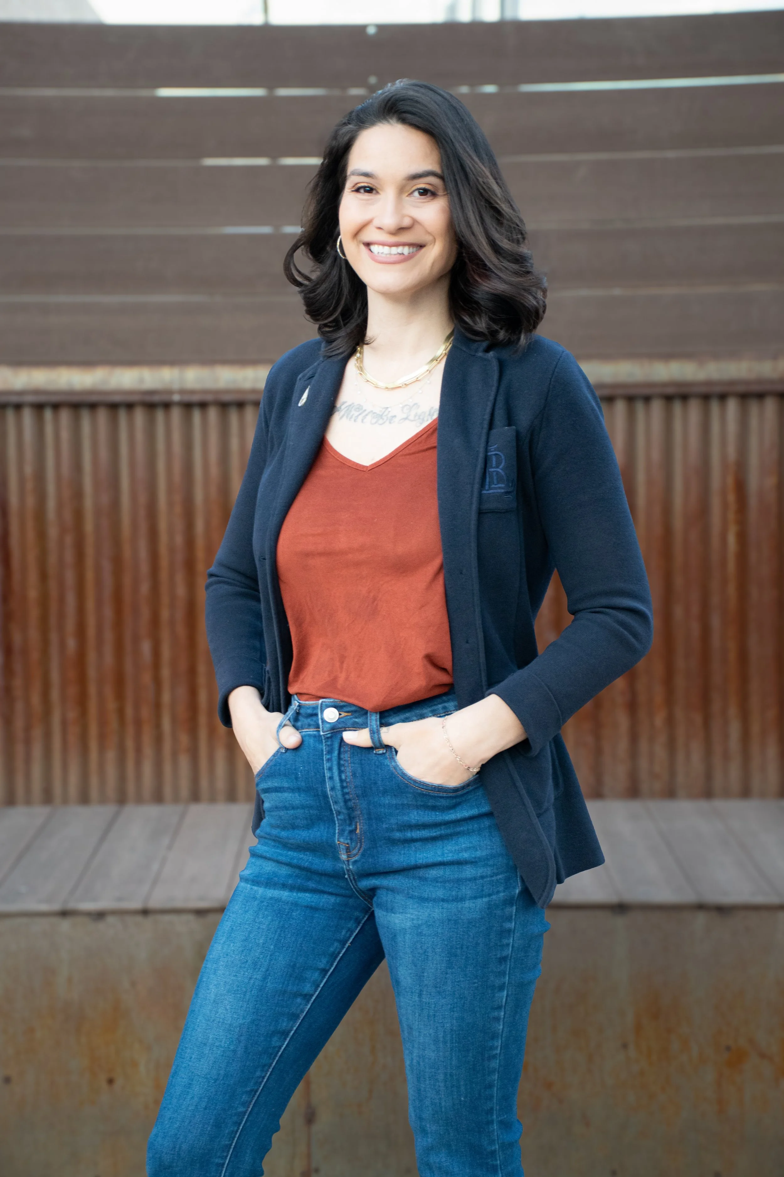 A woman with shoulder-length dark hair, smiling and standing outdoors in front of a wooden fence, wearing a navy blazer, rust-colored top, and blue jeans.