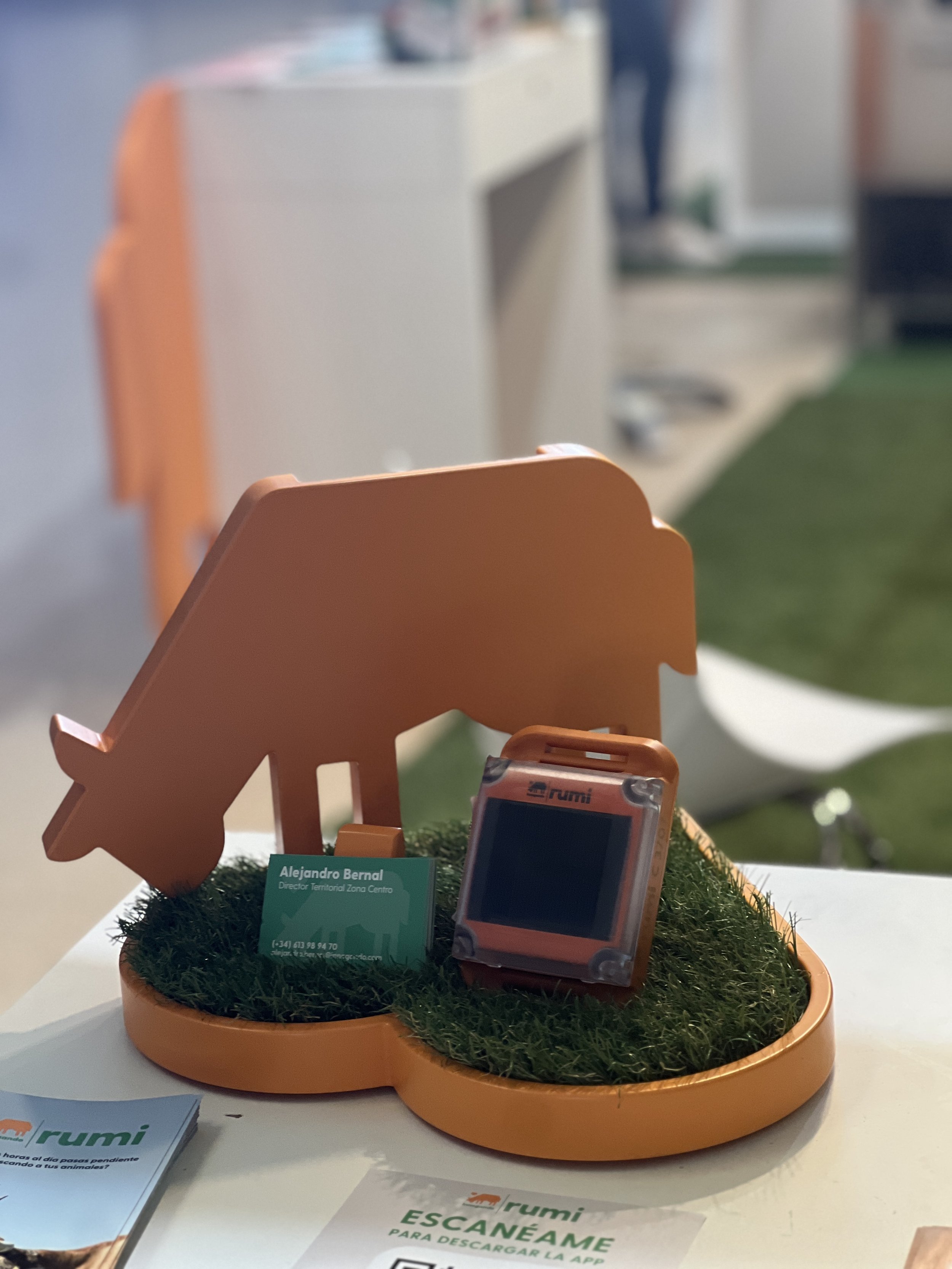 Muñeco de Minecraft en forma de zorro de color naranja, exhibido en una base de césped artificial en una mesa. Incluye una tarjeta de presentación con nombre y contacto, y material promocional de la empresa Rumi.