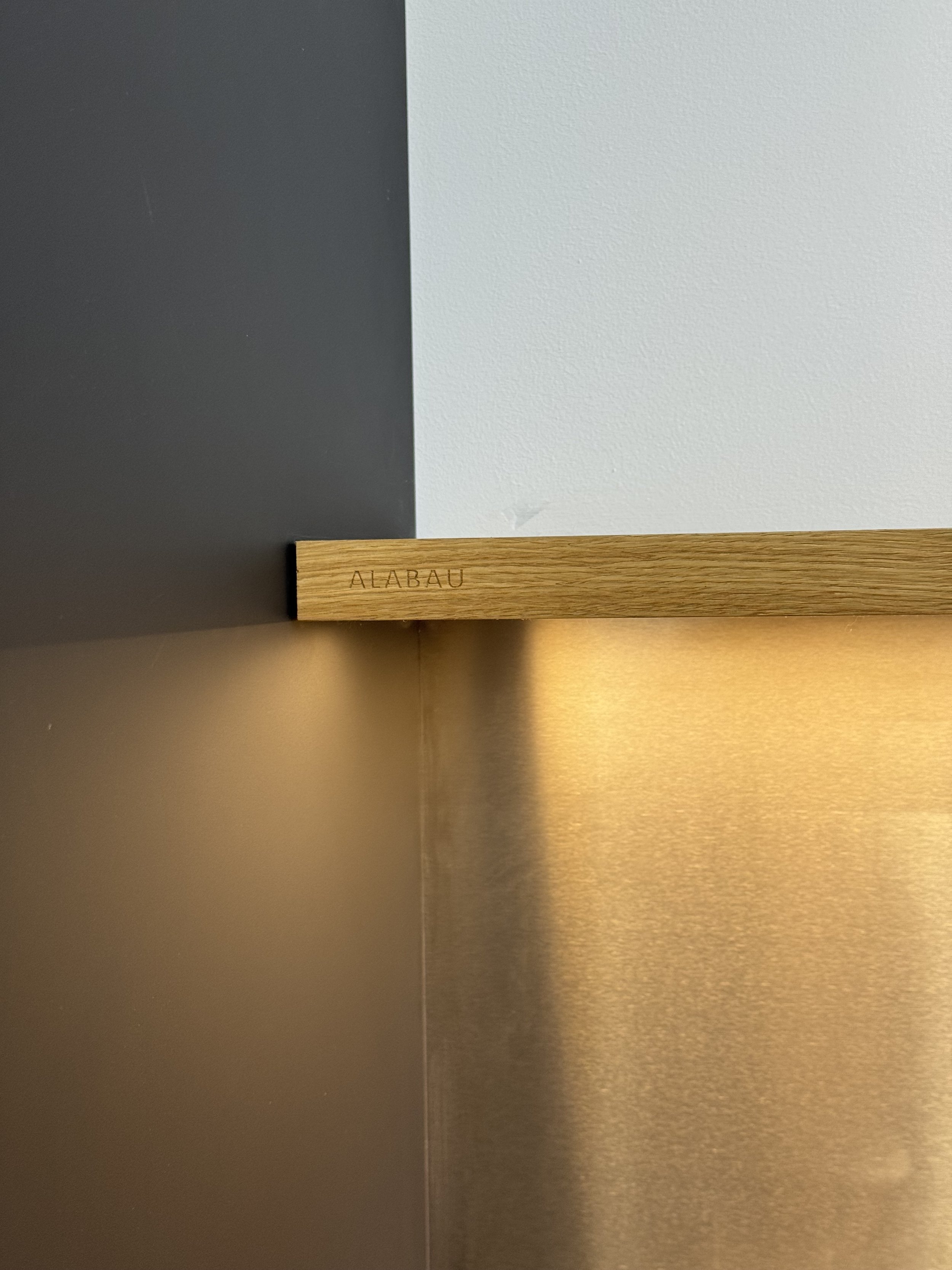 Madera con la palabra 'ALABAU' grabada en ella, colocada en una esquina de la pared, con colores gris oscuro, gris claro y dorado en el fondo.
