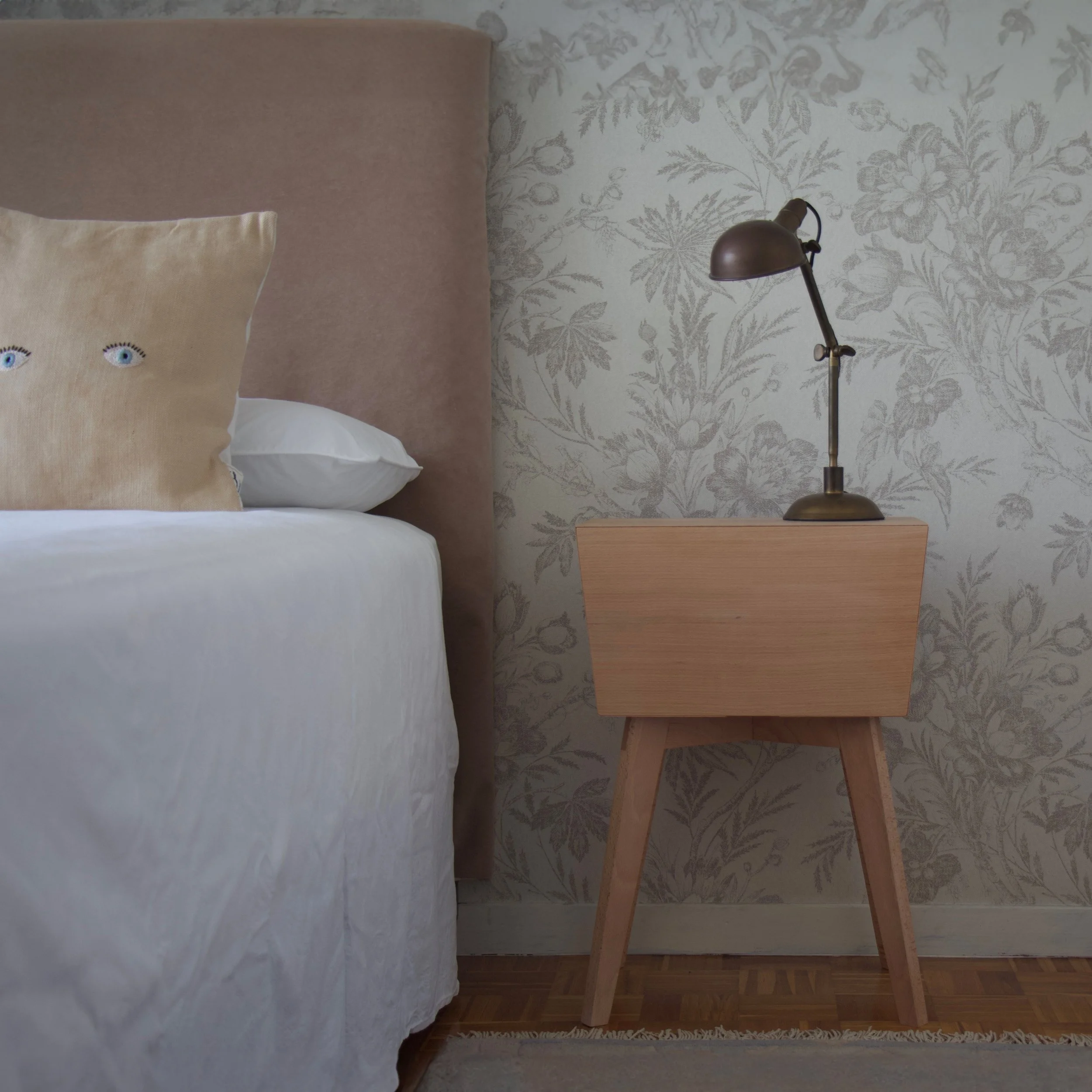 Imagen de una mesita de noche de madera con un lámpara de escritorio en la esquina, al lado de una cama con cabecero de color beige y una almohada con ojos bordados, en una habitación con papel tapiz de flores en tonos suaves.