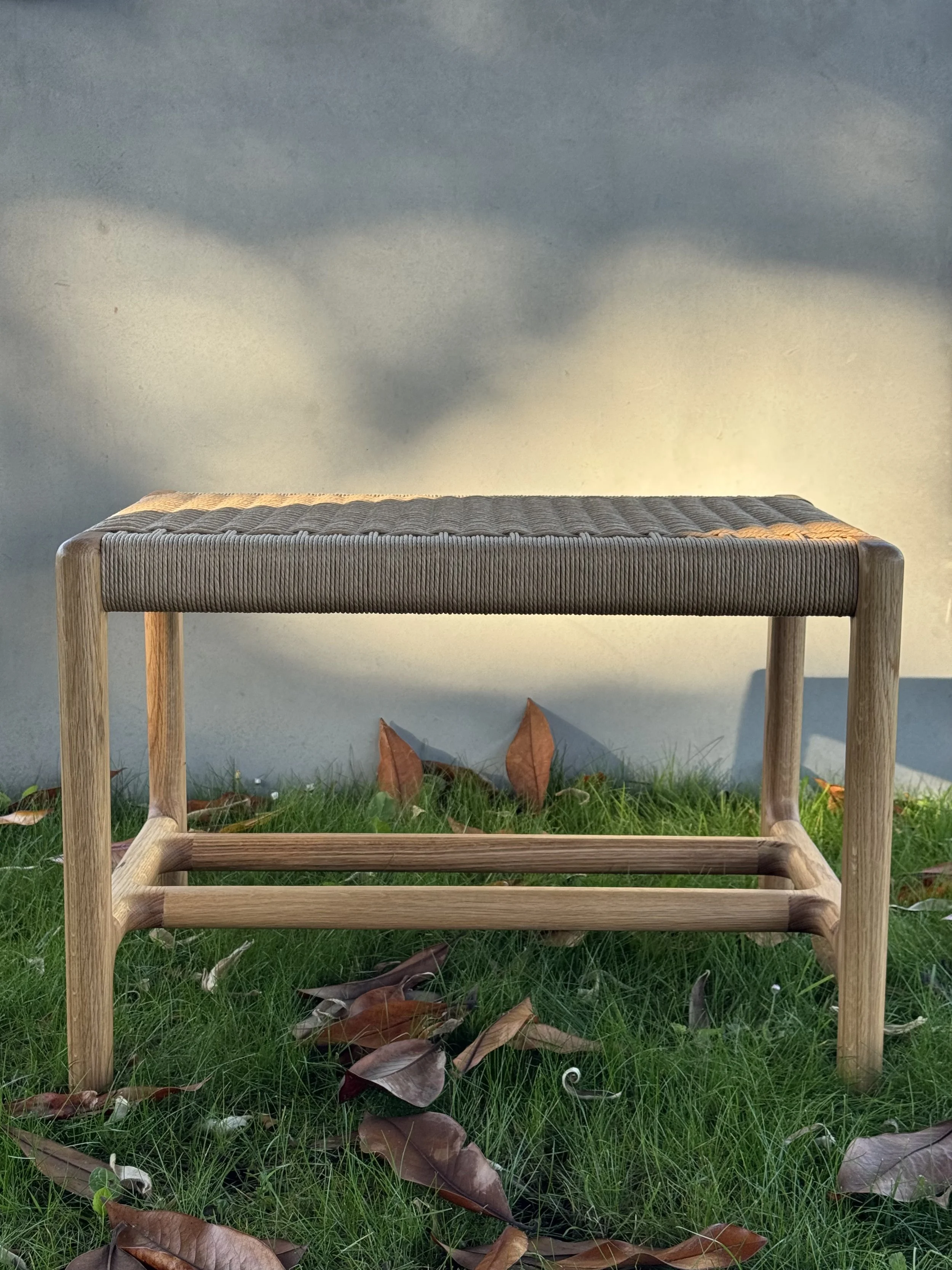 Banco de madera con asiento tapizado en la parte superior, sobre césped con hojas secas. Fondo de muro gris con sombras suaves.