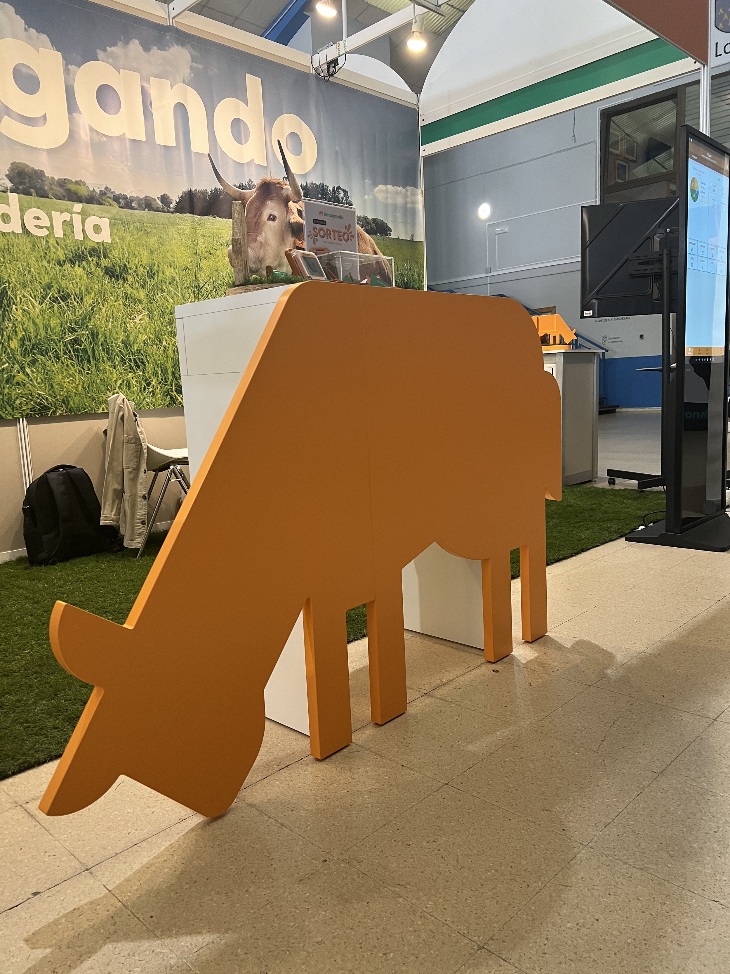 Una feria agrícola con un cartel que dice 'mercado' y una vaca grande en el fondo, junto a una mesa con una caja y algunos objetos. Hay una estructura de exhibición y una pantalla digital a la derecha.