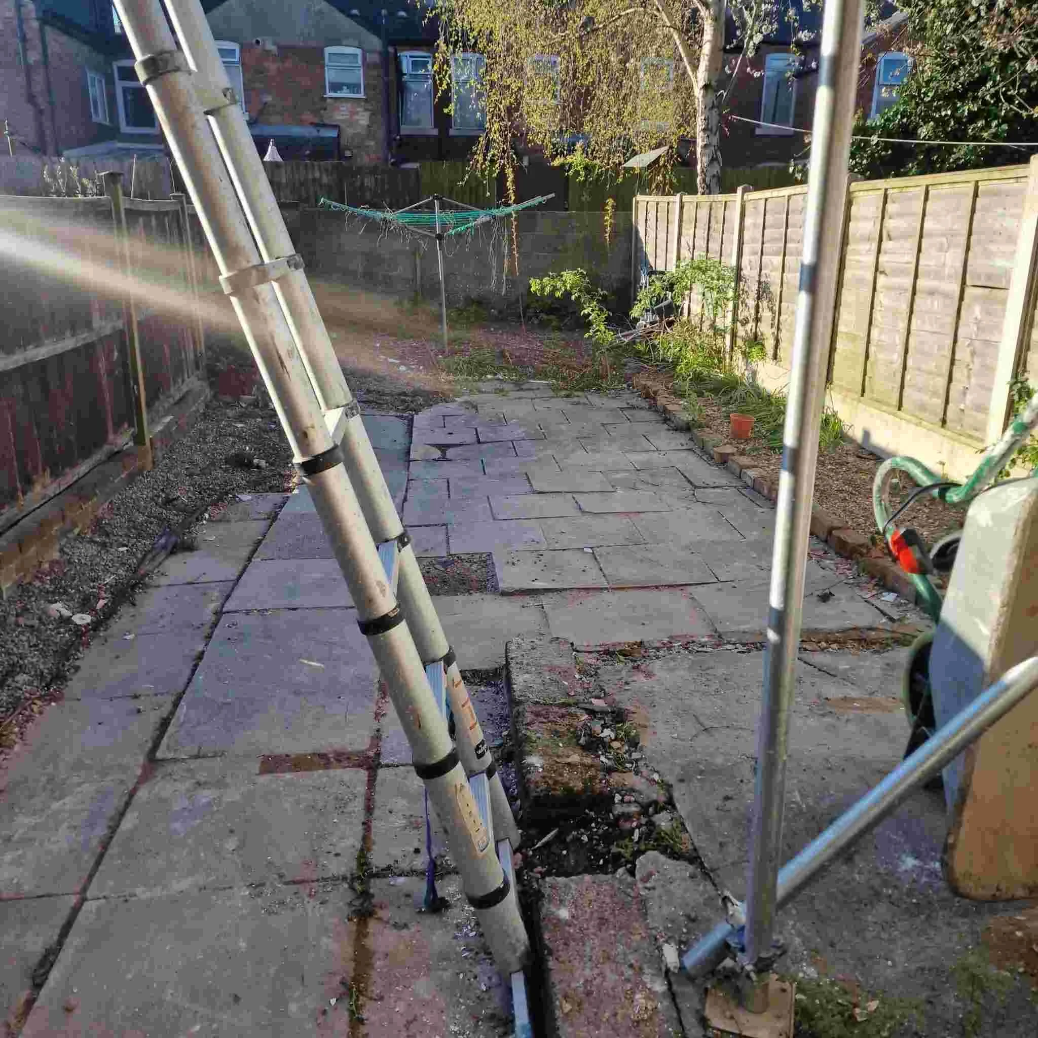A backyard with a paved patio area, a wooden fence, some trees, a clothesline with laundry, and gardening tools and plants.