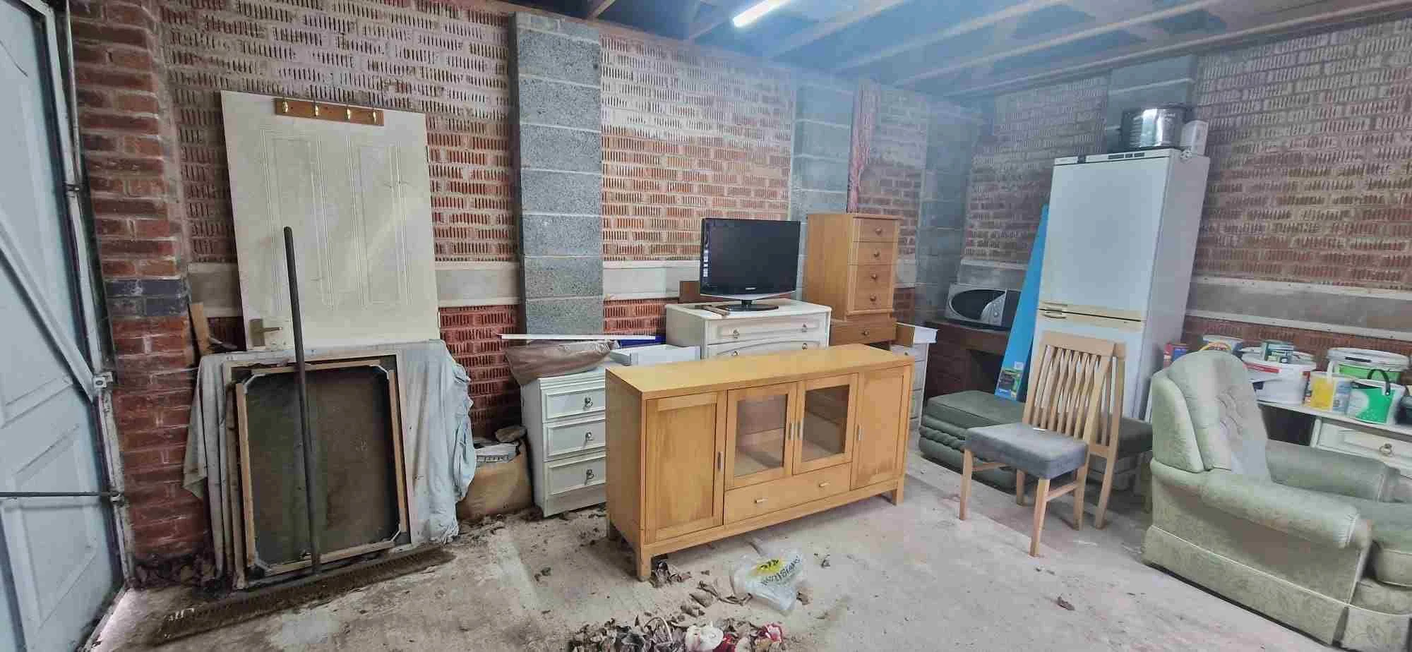 A cluttered room with brick walls, a small TV on a white dresser, a microwave, a refrigerator, dining chairs, a cushioned armchair, and various household items.