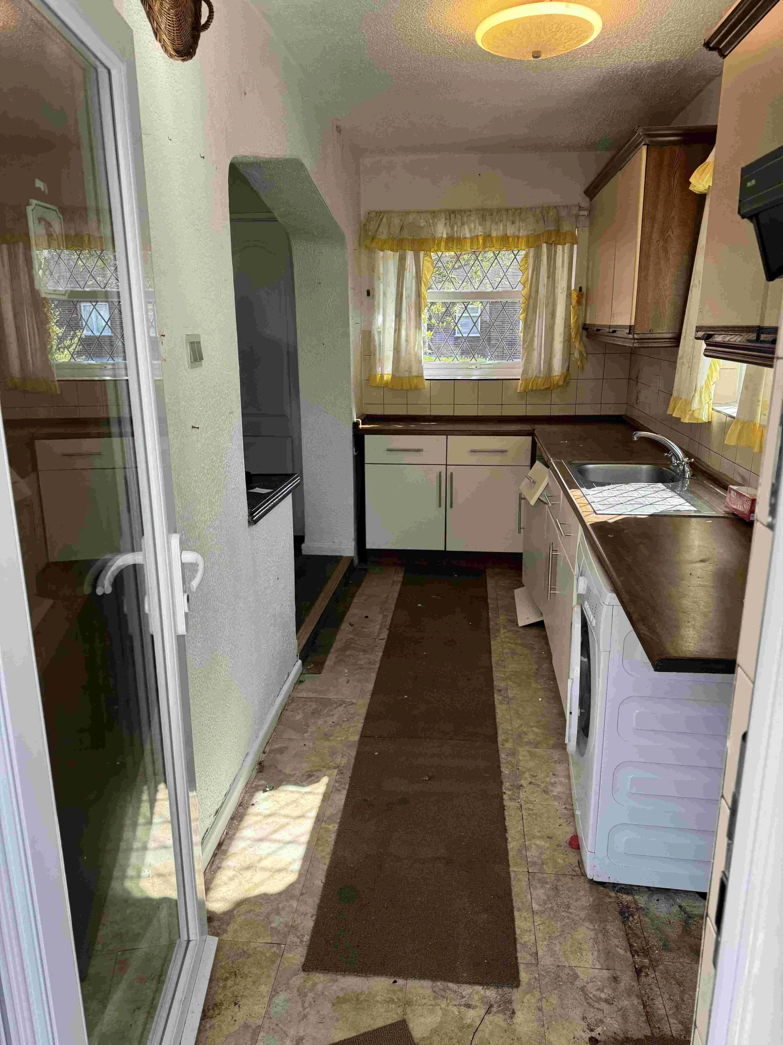 A narrow kitchen with a brown carpet runner along the floor, a window with yellow curtains, a kitchen sink, and white cabinets. There's a washing machine at the far right and a refrigerator at the far left. The flooring is tile, and the ceiling has a beige textured finish with a ceiling light.