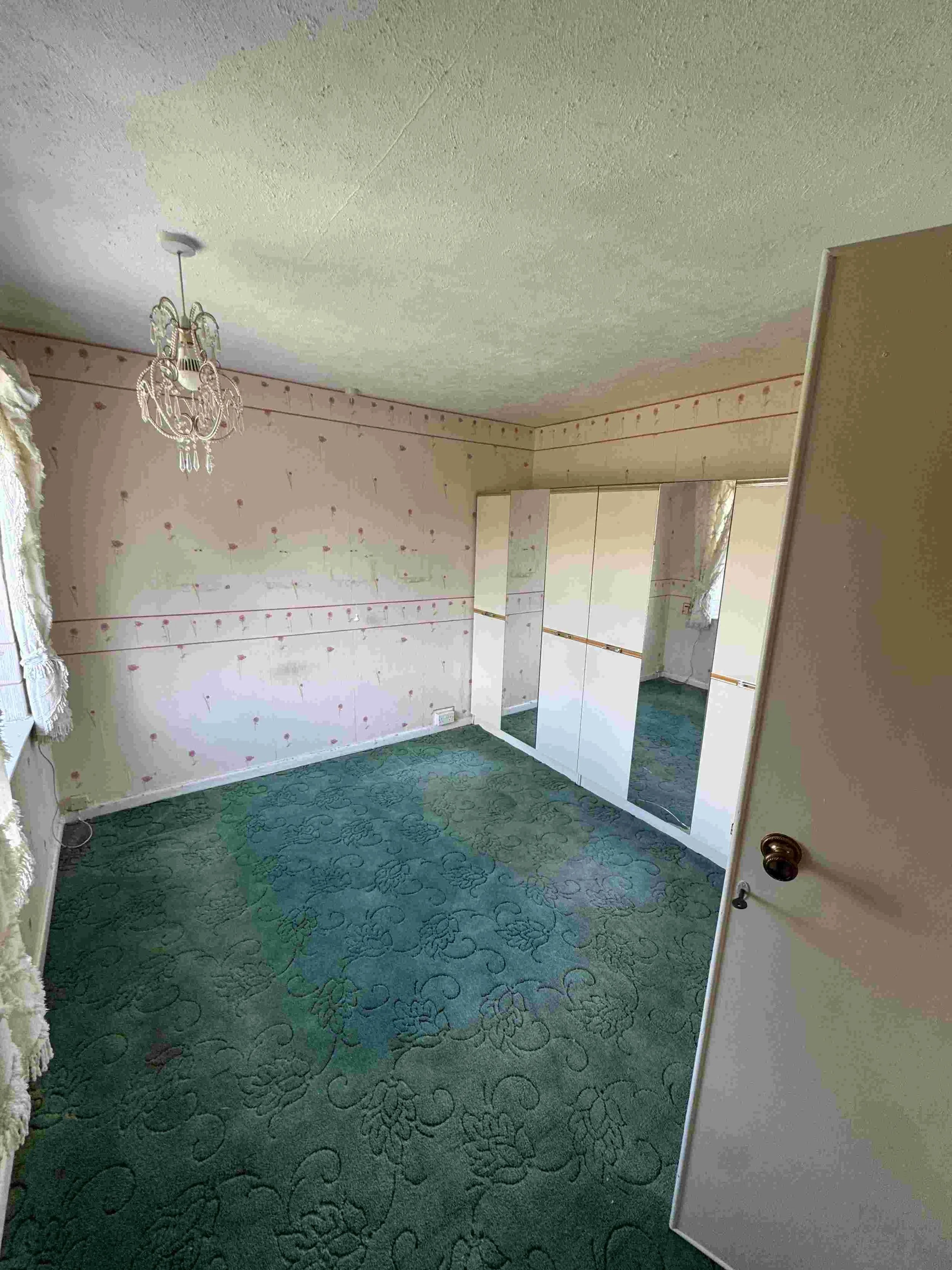 Empty room with green patterned carpet, floral wallpaper, white and gold closet, chandelier, window with lace curtains, and partially open door.