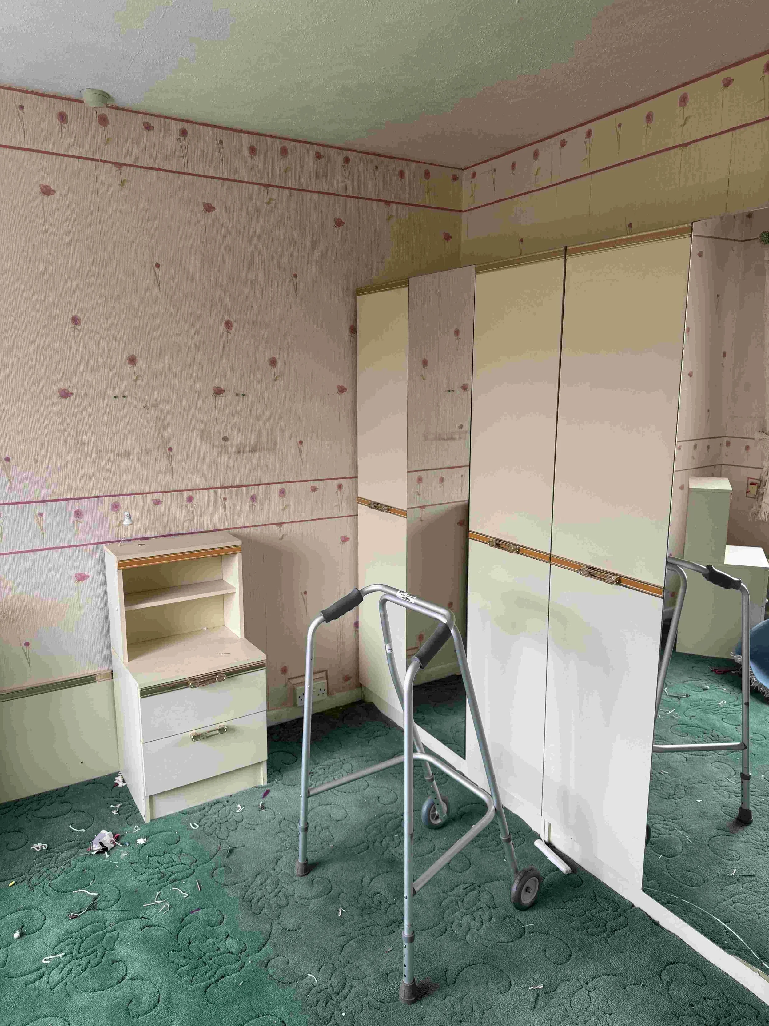 A room with floral wallpaper, a small white shelf, and a large mirror. There are two walkers in the room, and debris on a green patterned carpet.