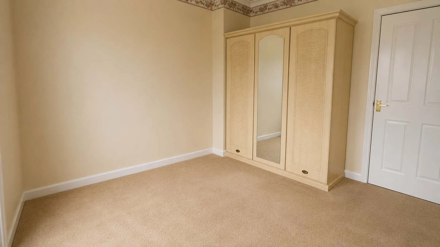 Empty beige room with carpeted floor, cream-colored walls, a wooden wardrobe with a mirror, and a white door.