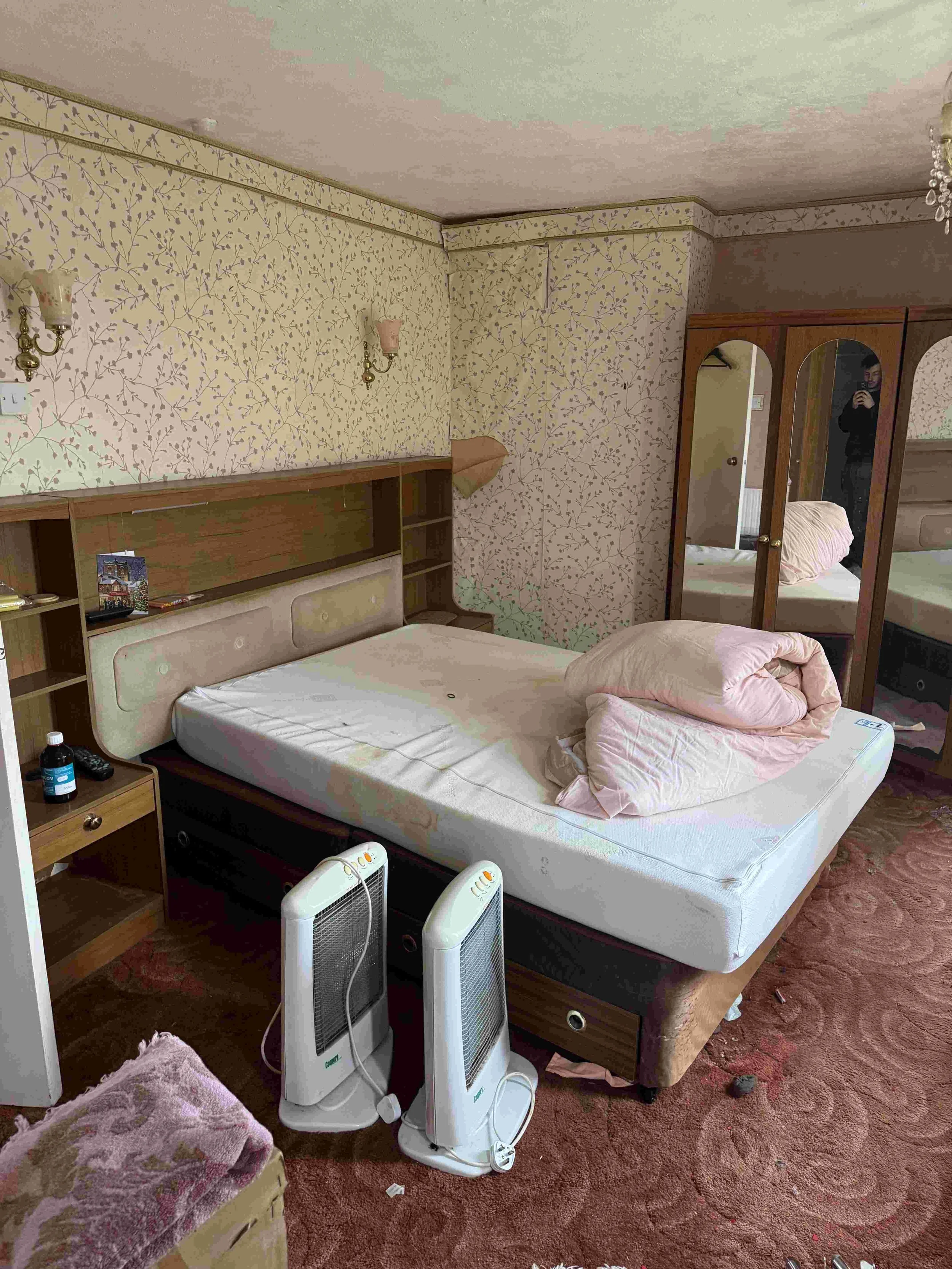 A messy bedroom with a unmade bed, two space heaters, and a brown carpet. There are small bedside tables, a mirror closet, wall-mounted lamps, and wallpaper with a floral pattern.