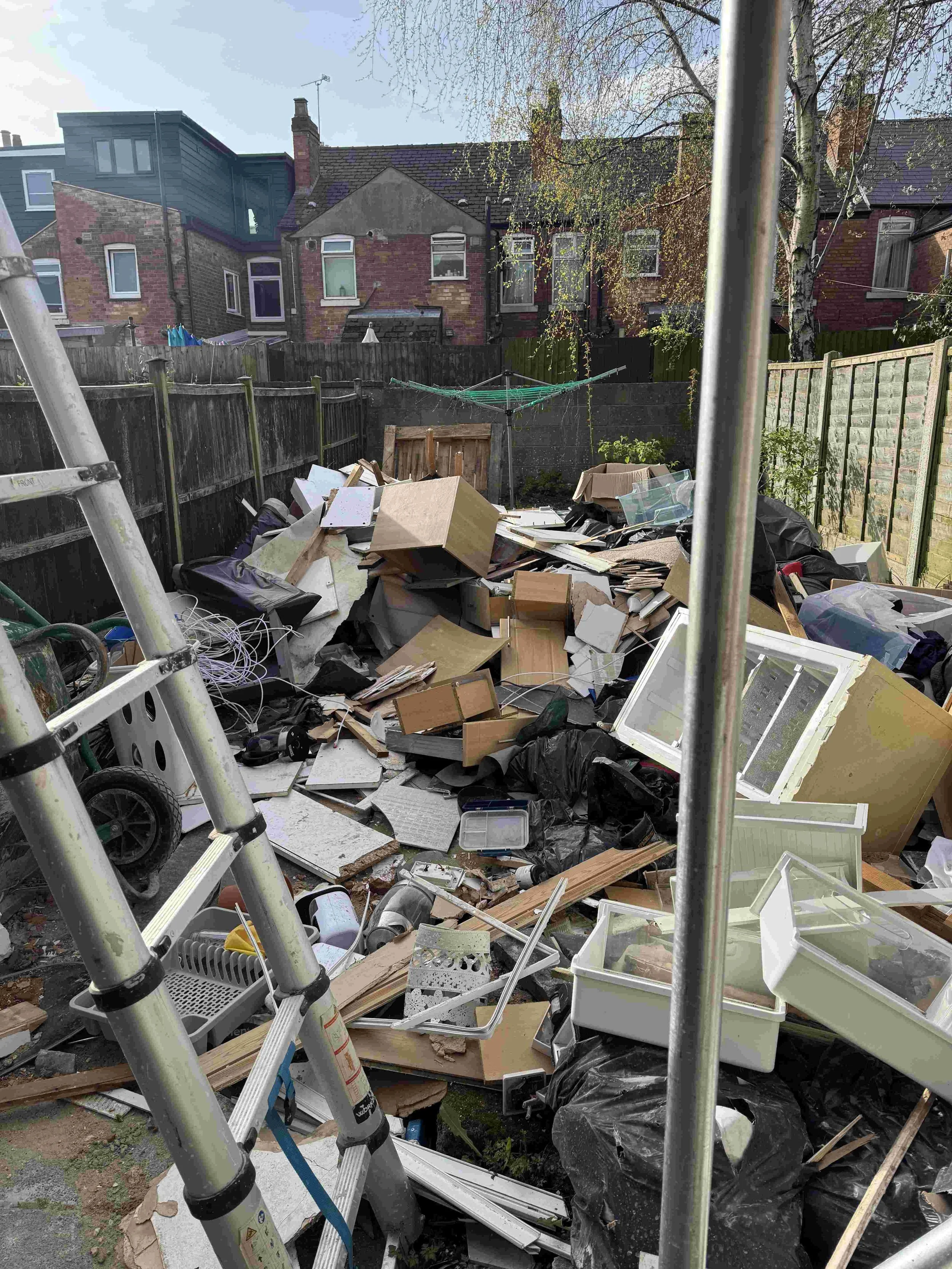 A backyard filled with assorted trash, including wooden planks, cardboard boxes, plastic containers, broken furniture, black trash bags, and debris. There is a metal fence, a ladder in the foreground, and a clothesline with clothespins. In the background, neighboring houses with brick walls and small windows are visible, along with trees and a partly cloudy sky.