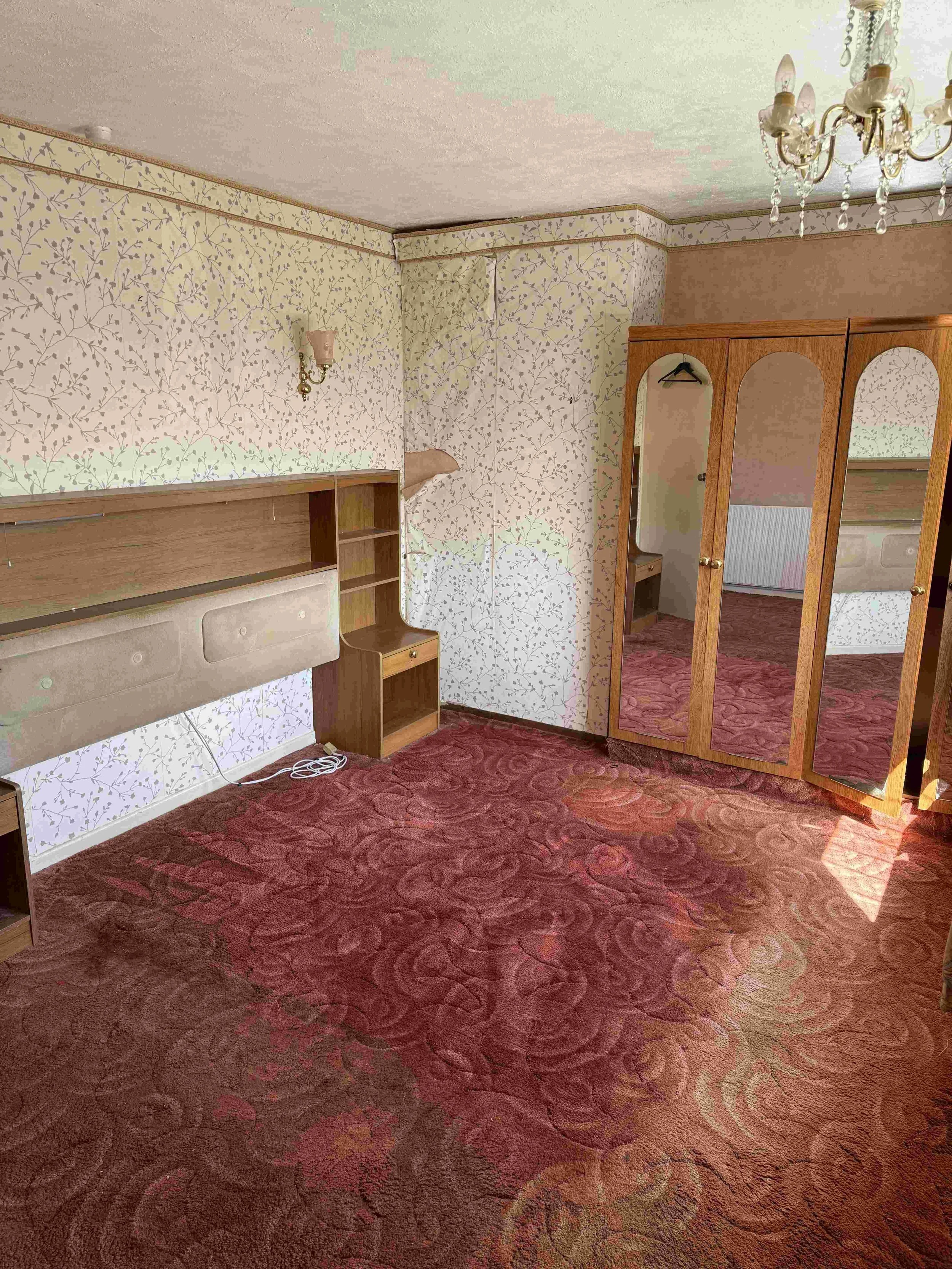 A bedroom with floral wallpaper on cream background, a wooden wardrobe with mirror doors, a wall sconce, a chandelier, and a red patterned carpet.