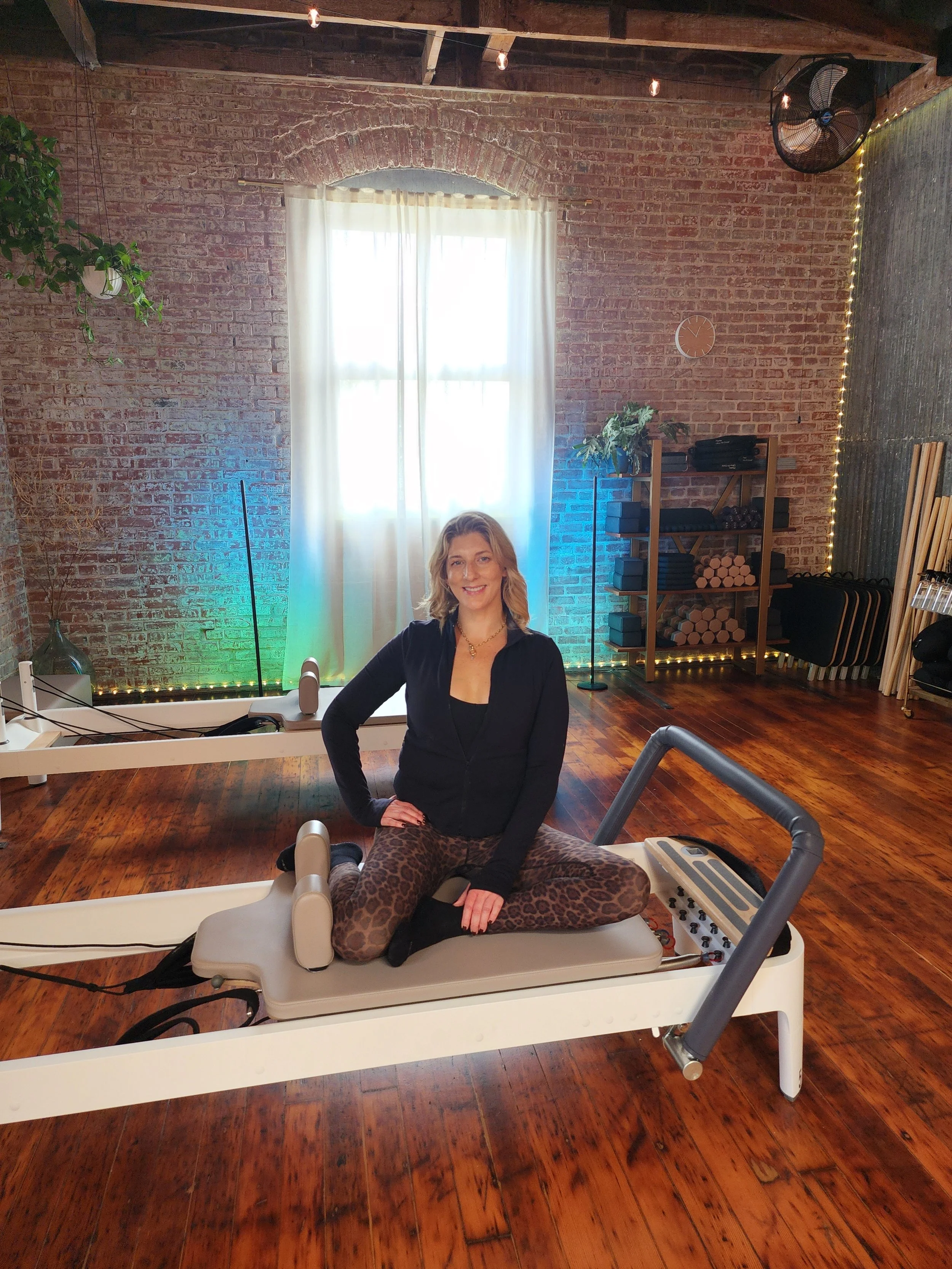 A woman in athletic wear is sitting on a Pilates reformer machine in a fitness studio with brick walls, hardwood flooring, and exercise equipment on shelves in the background.