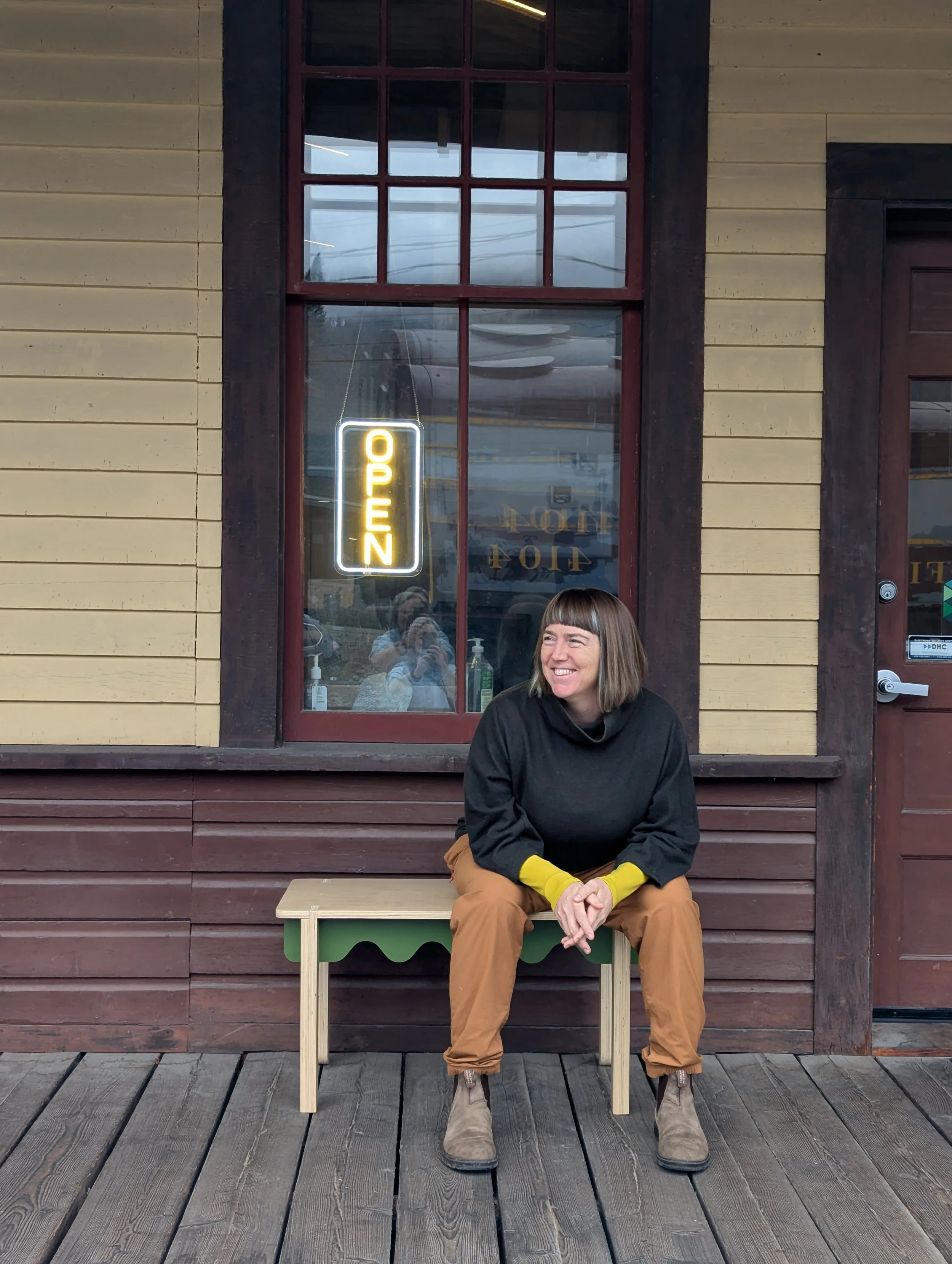 A woman with a bob haircut and bangs sitting on a small bench outside a building with a large window and a door. The woman is smiling and wearing a black top, brown pants, and beige boots. There is an illuminated sign in the window that says 'OPEN'.