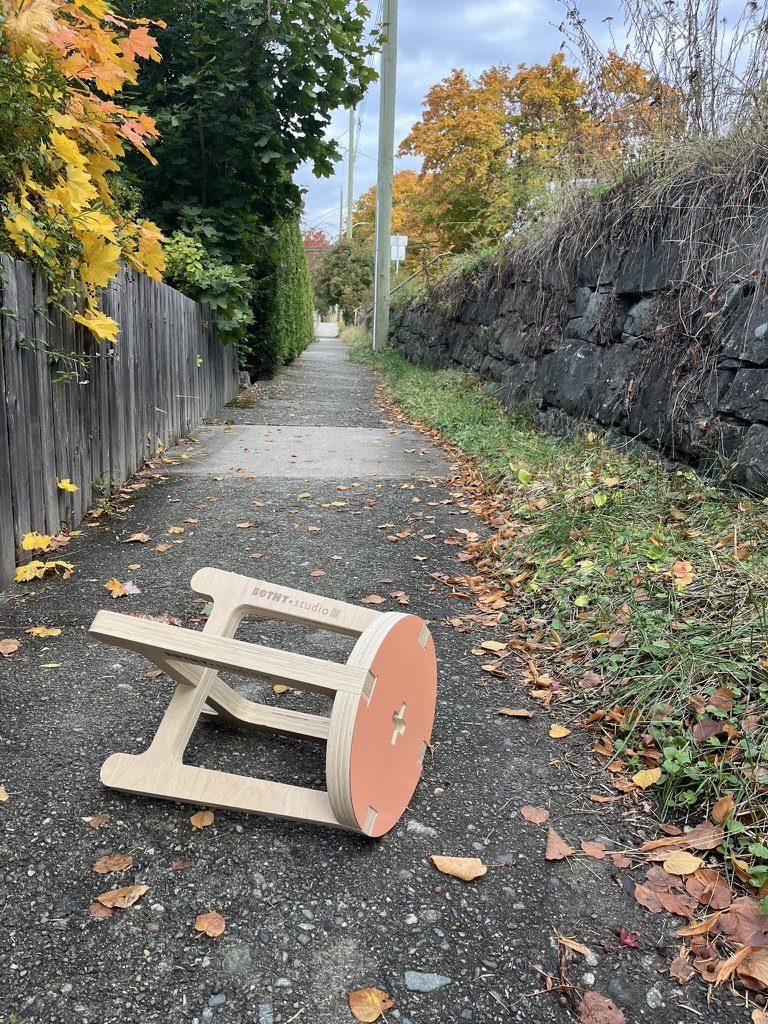 A small overturned wooden chair with a round orange backrest lying on a leaf-covered sidewalk in an outdoor setting.