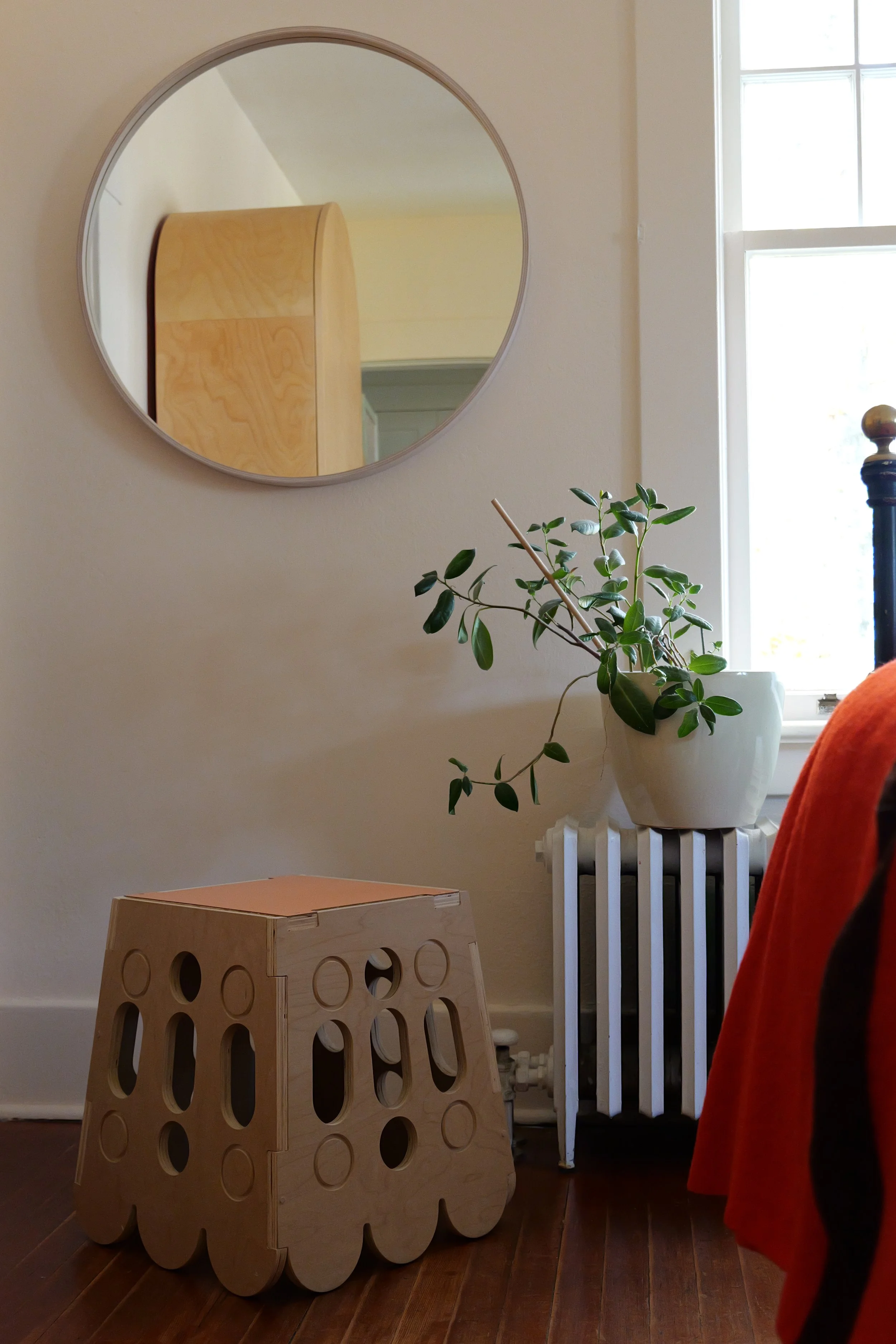 Interior scene showing a round mirror on the wall reflecting a wooden cabinet, a potted plant on a white radiator, a wooden stool with circular cutouts, part of a table with an orange tablecloth, and a window with sunlight.